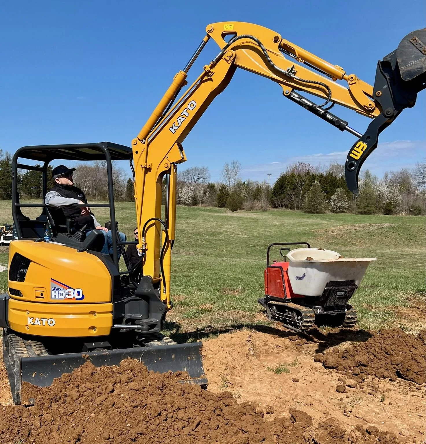 A man operating a yellow KATO excavator on grass, with a small tracked utility vehicle and a wheelbarrow nearby, under a clear blue sky.