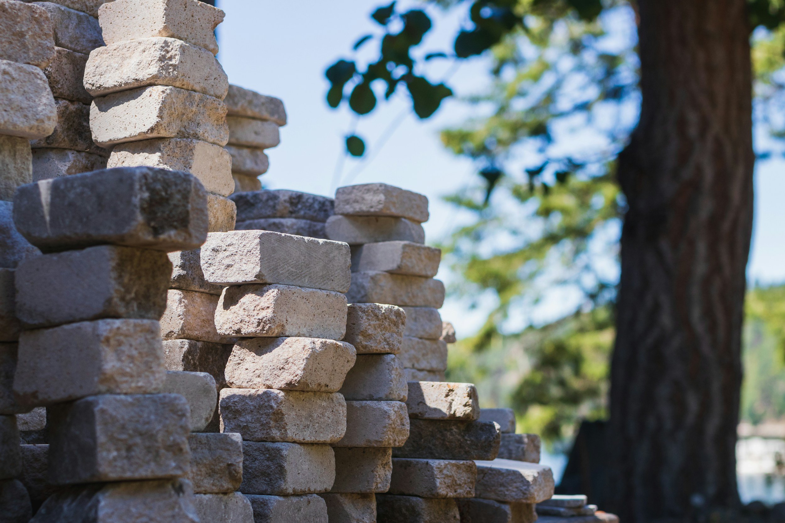 A stack of beige and gray stones arranged in a tower outdoors, with a background of green leaves and a tree trunk under a blue sky.