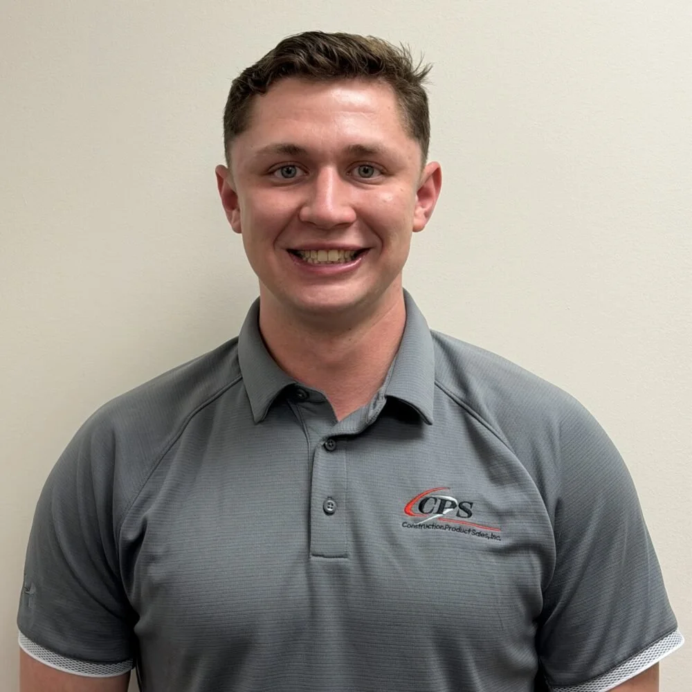 A smiling young man with short brown hair wearing a gray polo shirt with a logo that says 'CPS' standing against a plain beige wall.