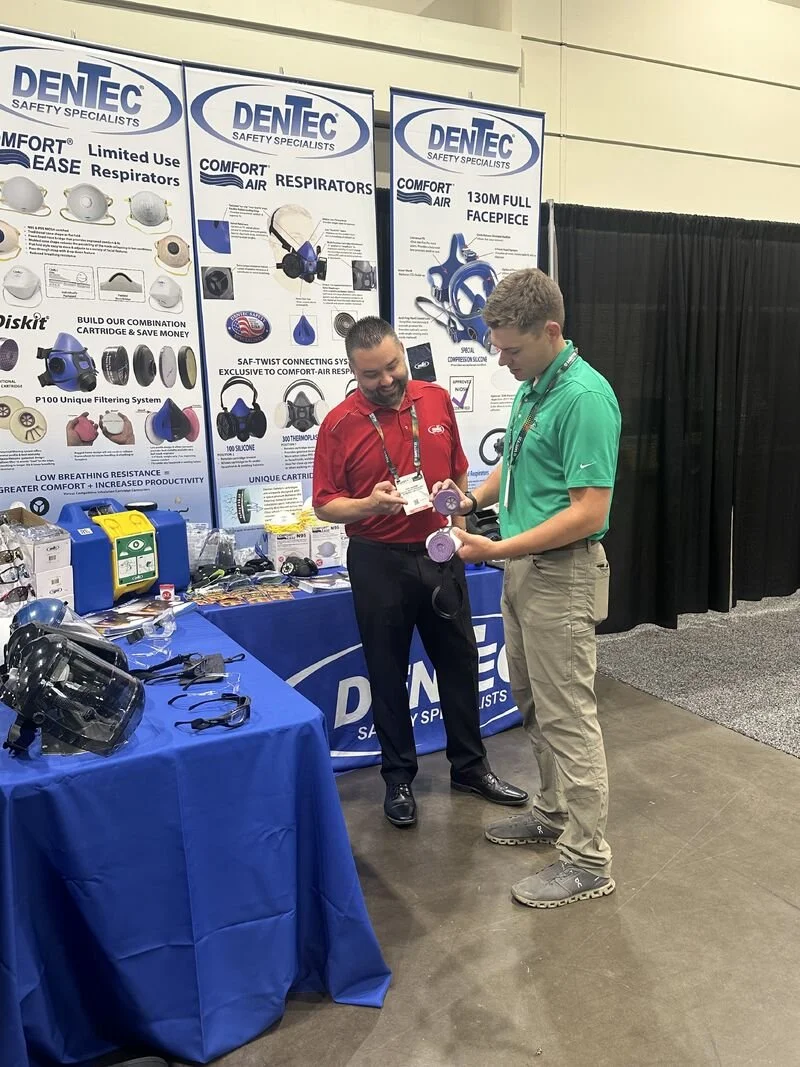 Two men at a DENTEC booth at a trade show. One man in a red shirt is showing a purple mask to the other man in a green shirt. The booth displays various respiratory and safety products, with large banners featuring product images and descriptions.