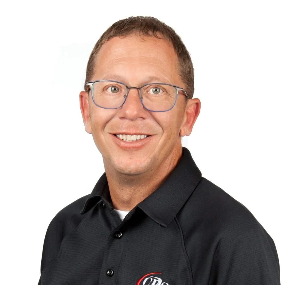 Portrait of a man with short brown hair, glasses, wearing a black collared shirt with logo, smiling against a white background.
