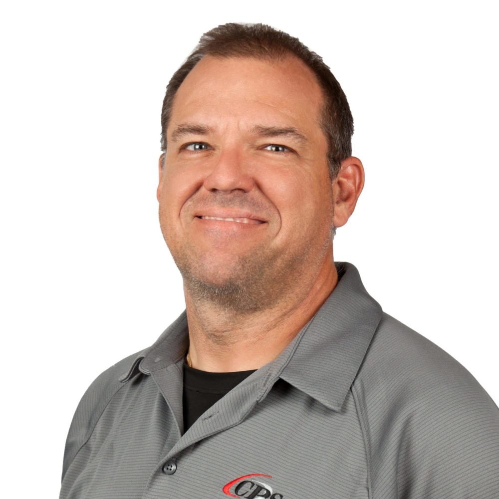 A man with short dark hair and a light beard wearing a gray collared shirt with a logo, smiling in front of a white background.