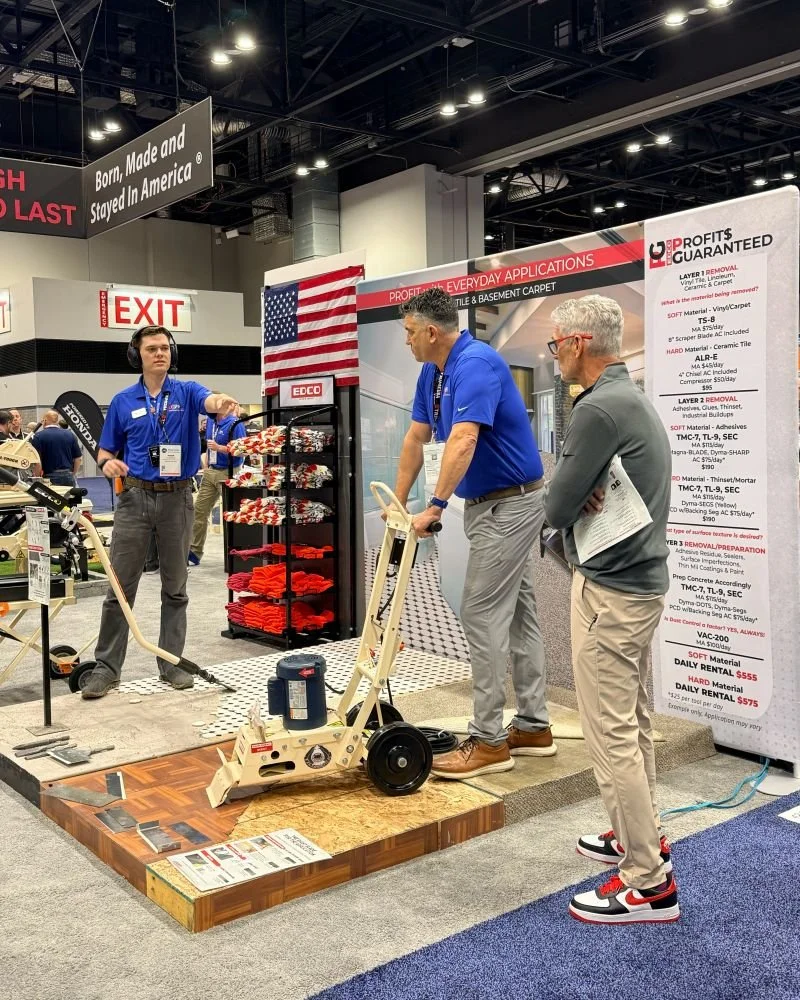 Two men and a woman demonstrating floor grinding equipment at a trade show booth. The booth features a sign with American flags and text. The booth is set up on a platform, with informational posters and product displays around.