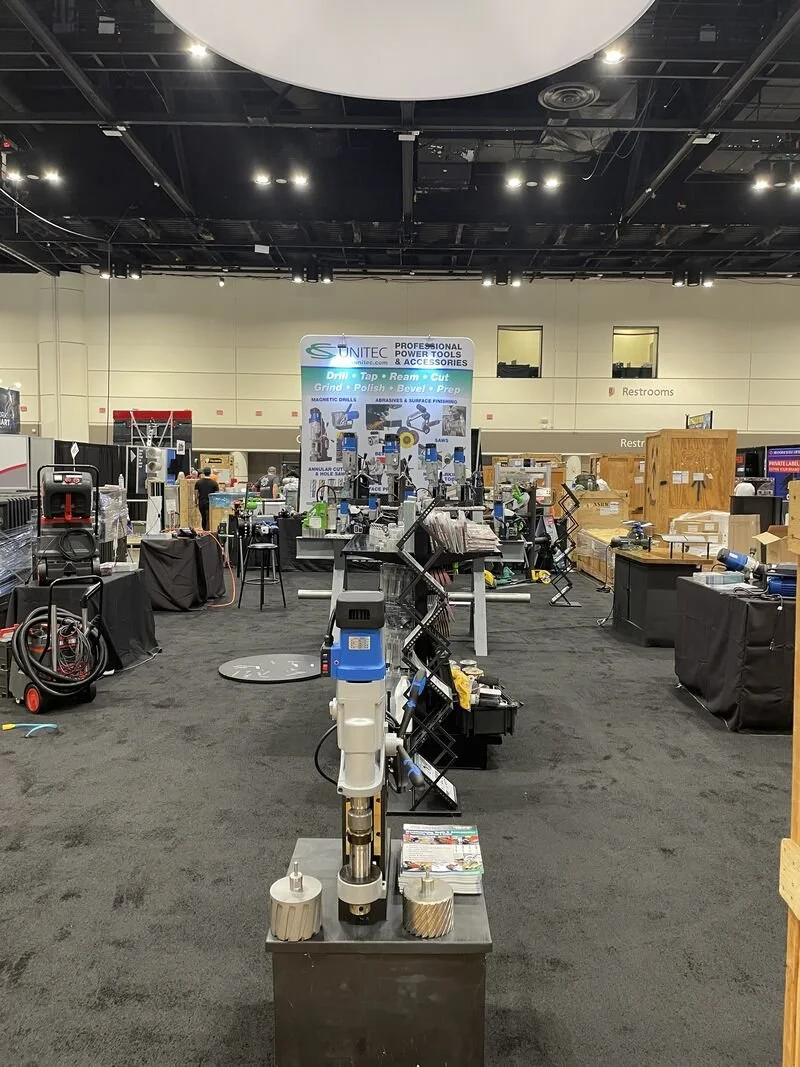 Trade show booth showcasing professional power tools and accessories, with various equipment, posters, and displays on black carpets in an exhibition hall.