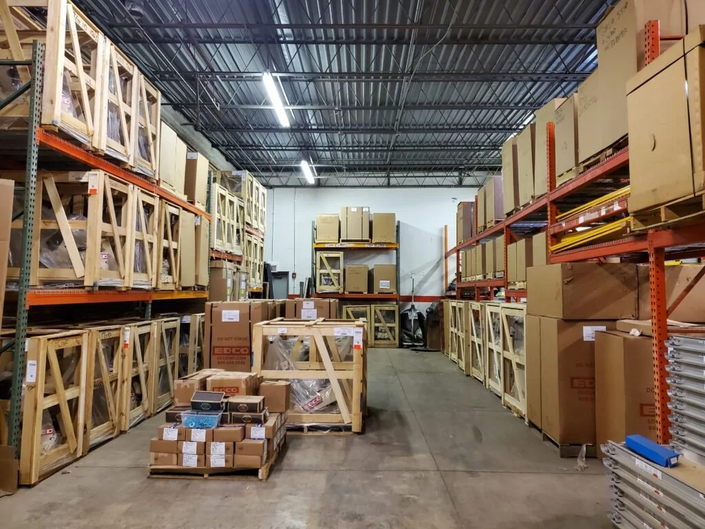 Warehouse aisle with shelves filled with cardboard boxes and wooden crates, some labeled, arranged on metal racks, with a concrete floor and metal ceiling.