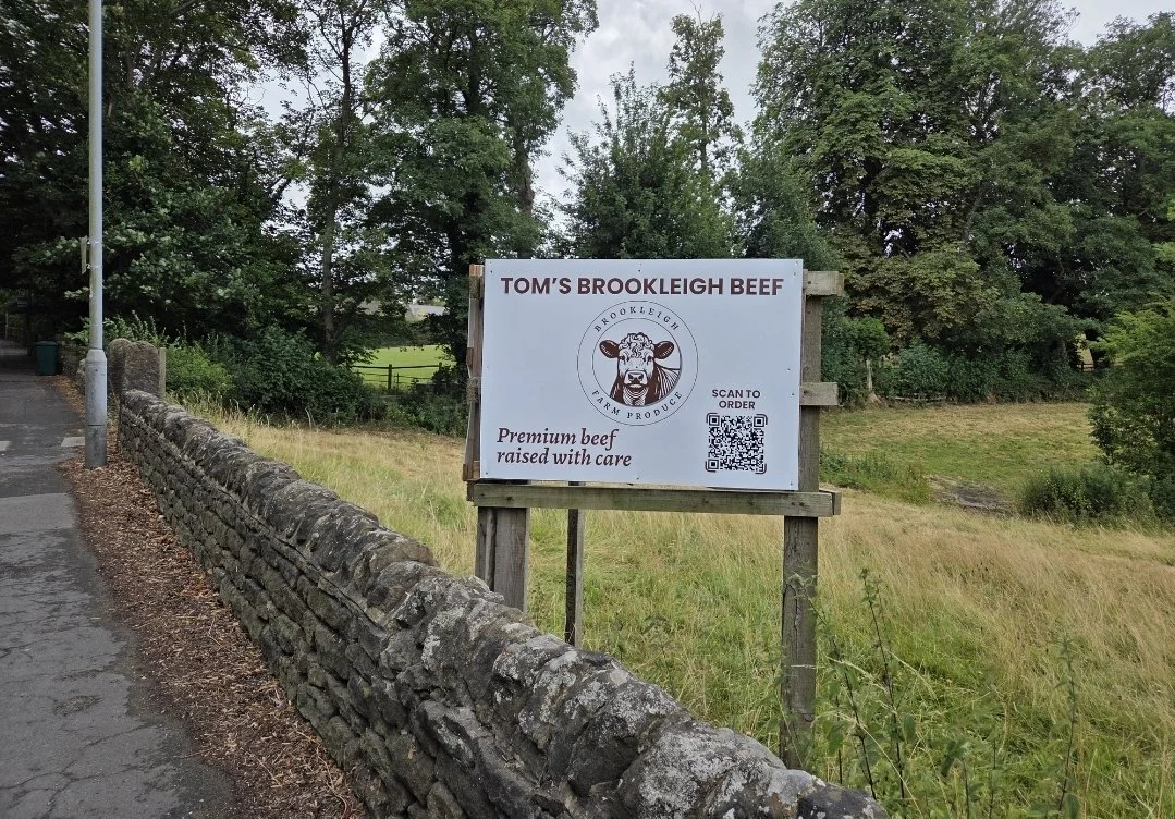 Signboard advertising Tom's Brookleigh Beef, premium beef raised with care, featuring a logo with a cow's head and QR code for ordering, located beside a stone wall and street, with trees and grassy field in the background.
