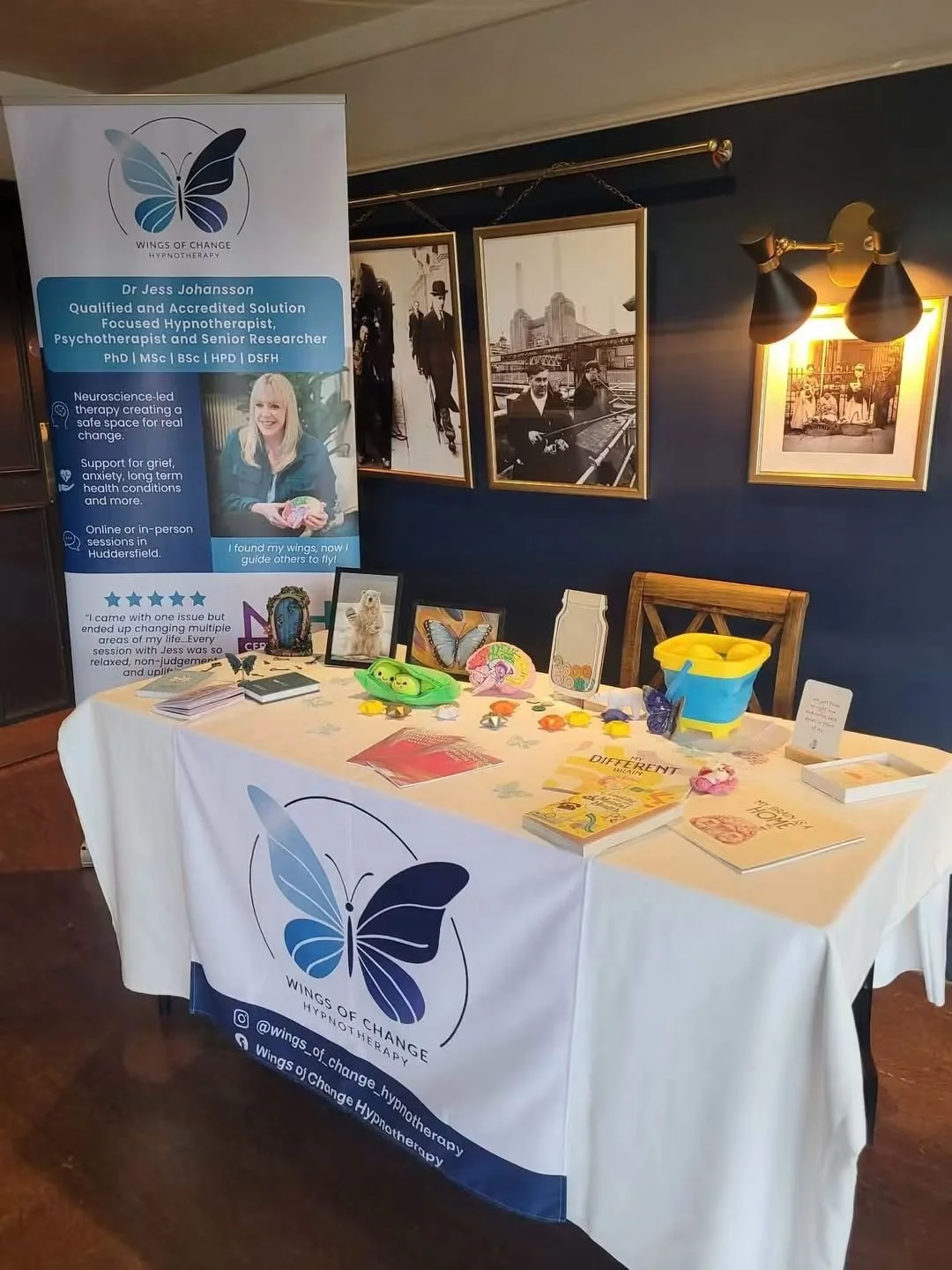 Display table with Wings of Change Hypnotherapy promotional materials, including a banner about Dr. Jess Johansson, framed photos on the wall, and various small items and brochures on the table.