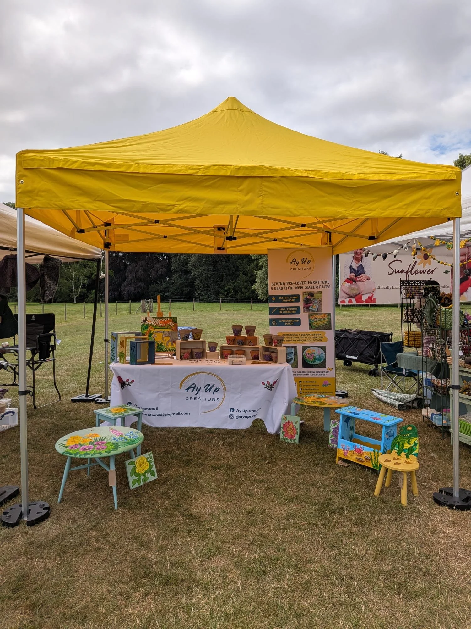 Outdoor booth with yellow canopy tent displaying painted wooden furniture and artwork, set on grass field under cloudy sky.