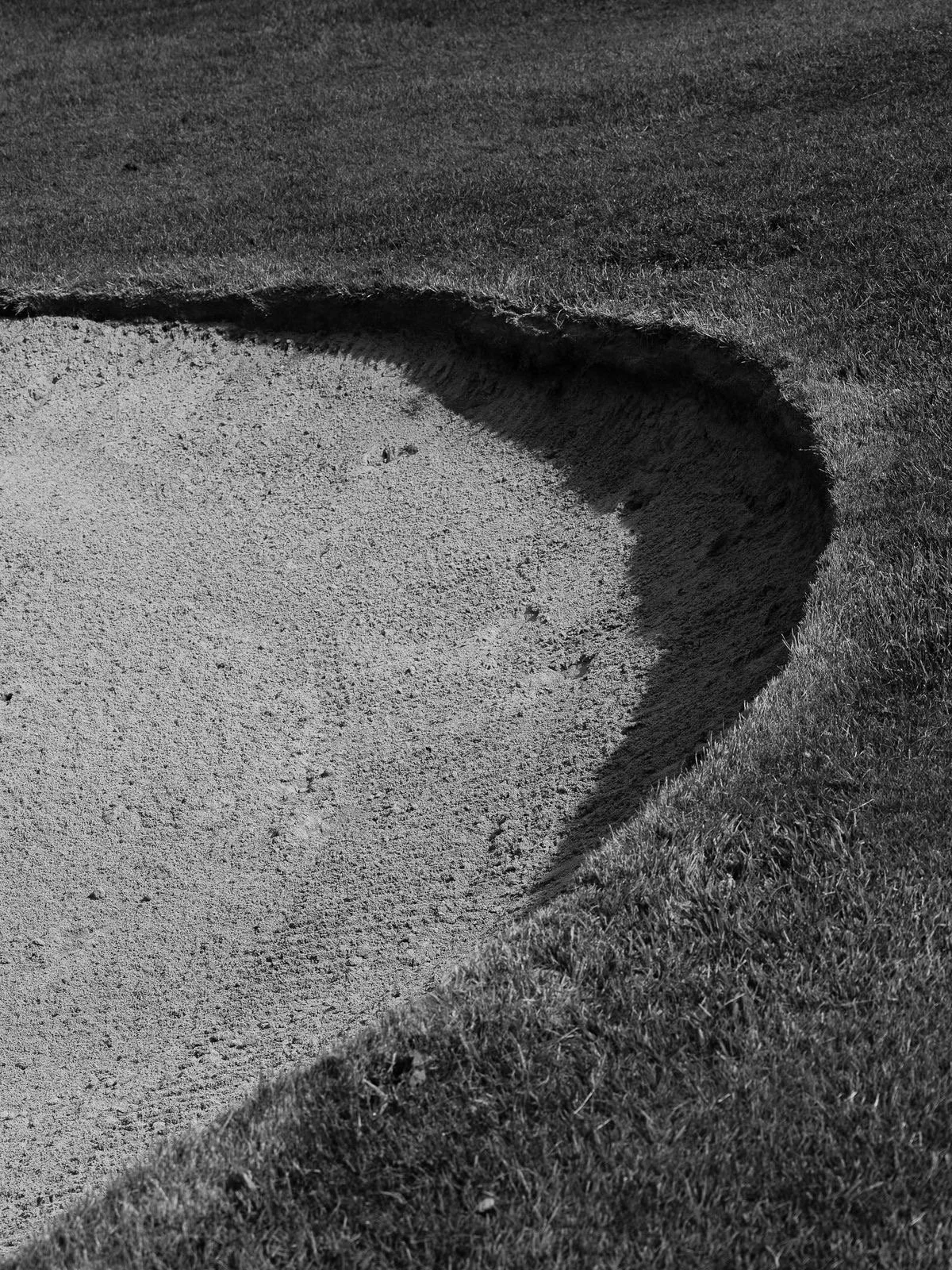 A golf bunker with sand and an edge of grass on a golf course, captured in black and white.
