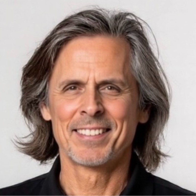 A middle-aged man with long, graying hair and a cheerful smile, wearing a black shirt, in front of a plain light background.