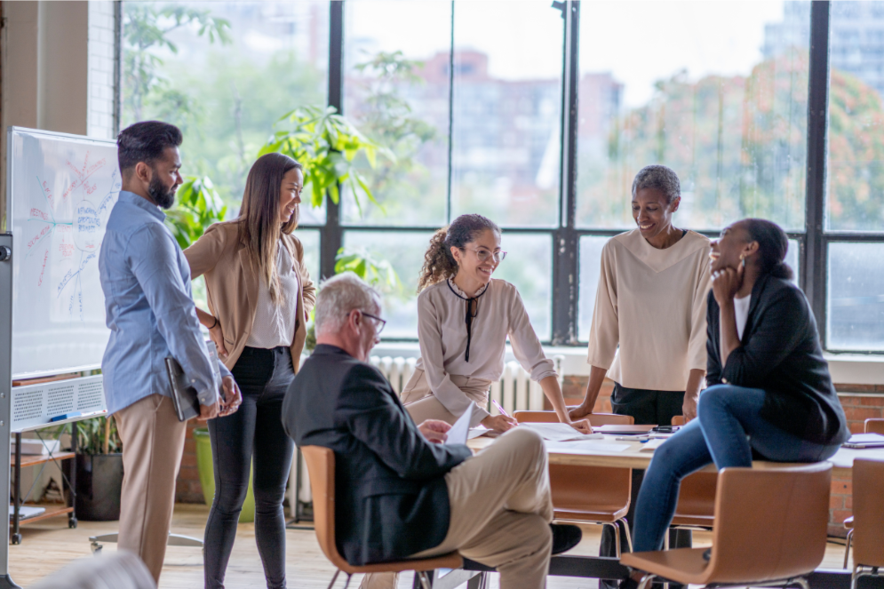 Group of six diverse professionals in a bright office space engaging in a discussion around a table, with large windows and greenery outside.