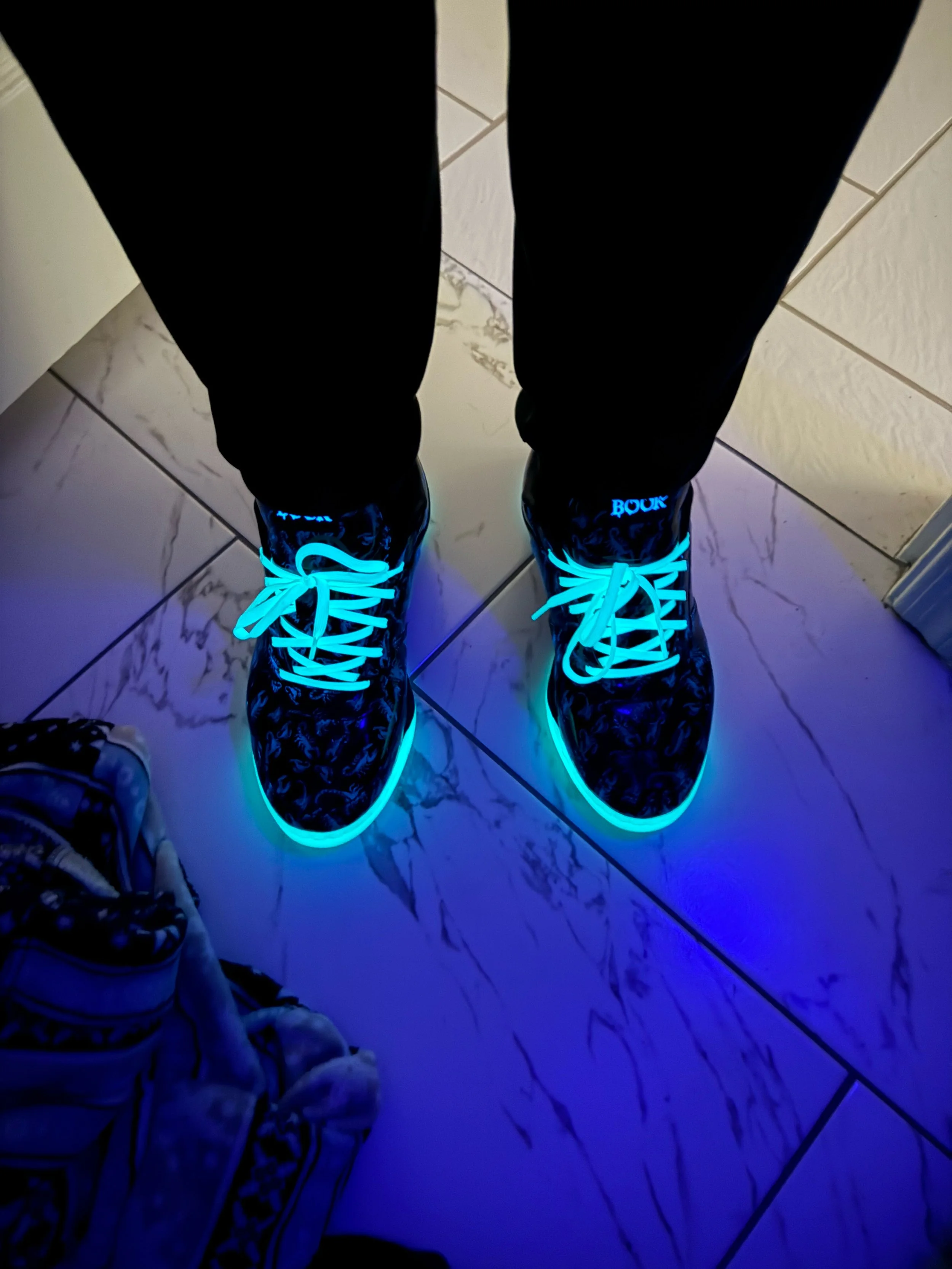Person standing on a tiled floor wearing black pants and sneakers with glowing blue soles and laces.