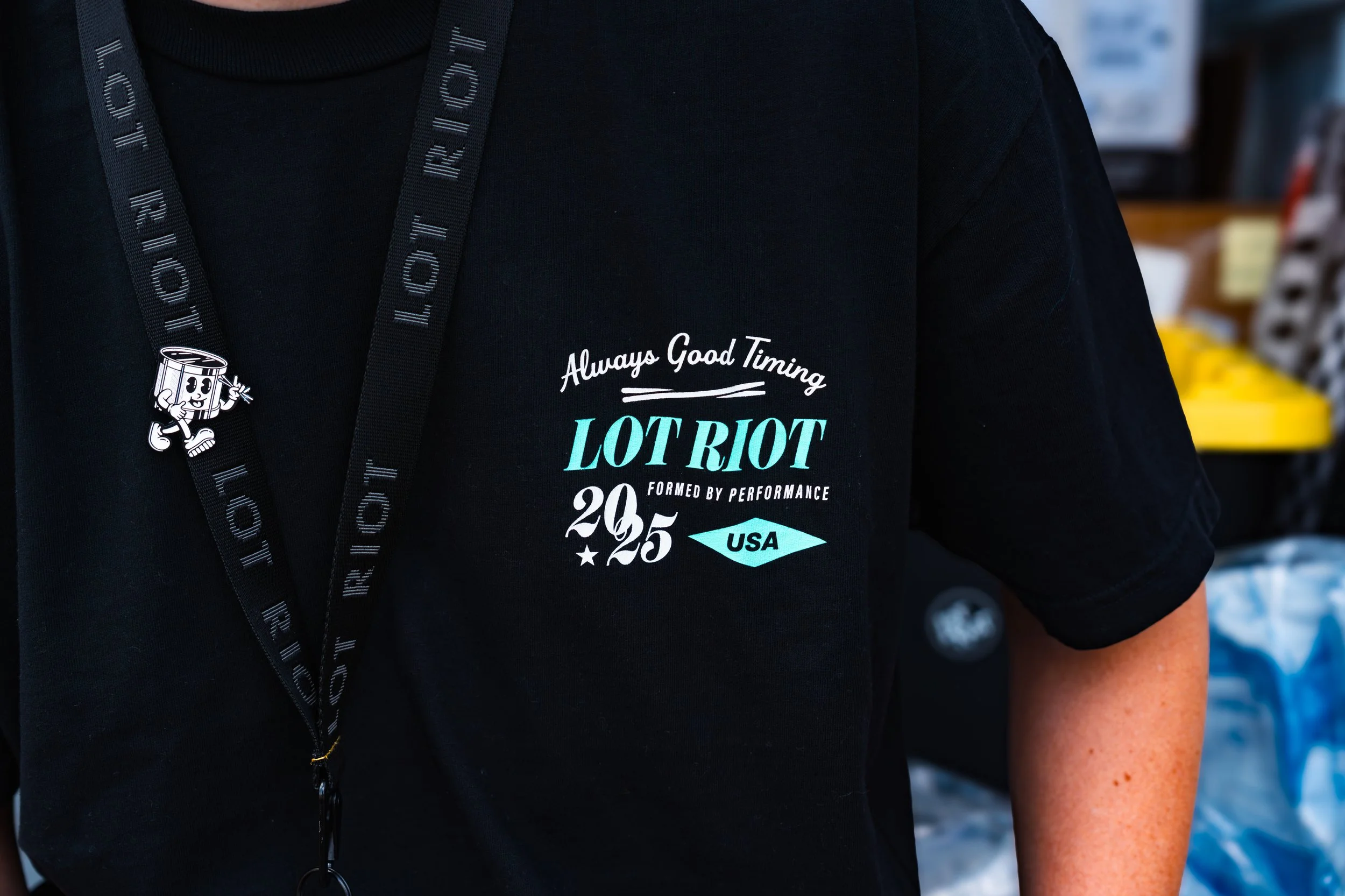 Close-up of a person wearing a black T-shirt with white and turquoise text and graphics, and a black lanyard with the words 'LOT RIOT' around their neck
