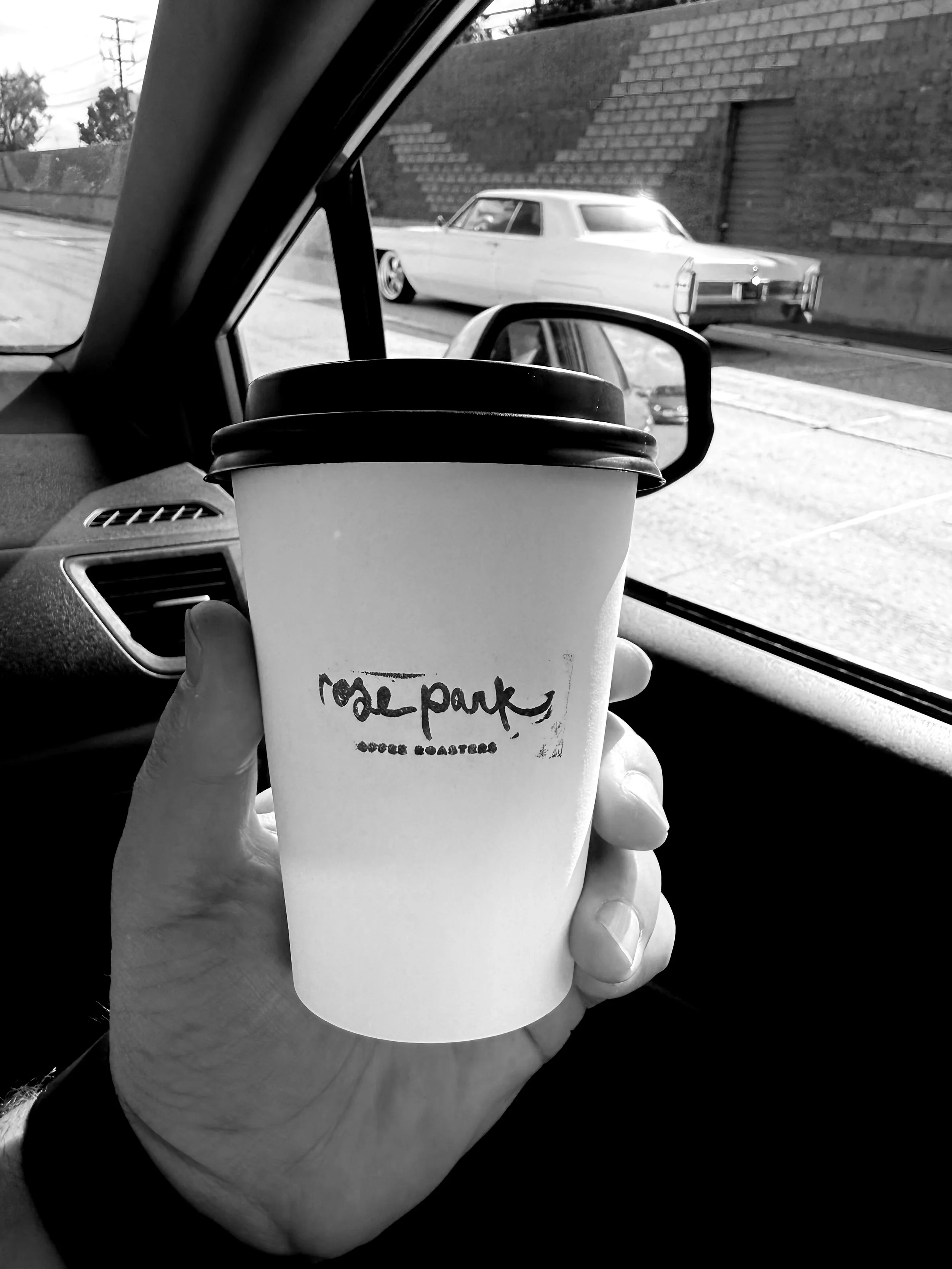 A person holding a disposable coffee cup with a black lid, inside a vehicle. The cup has handwritten text that reads 'rose park' and some smaller text below, with the background showing a street view with a vintage car passing by.
