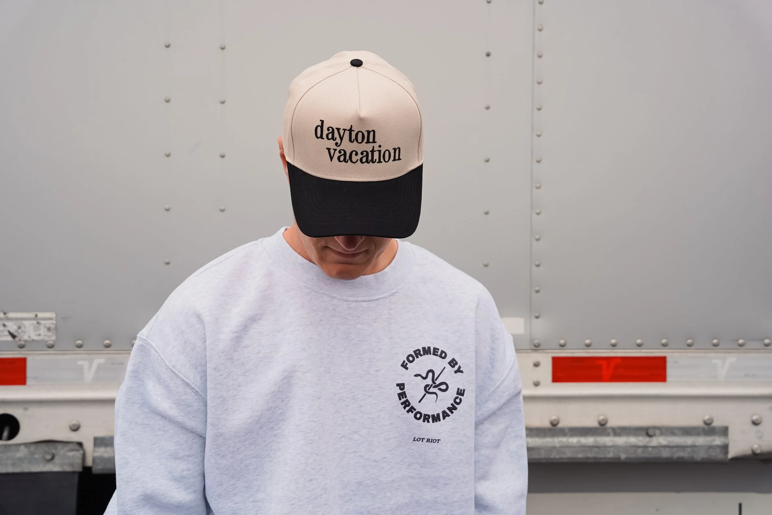 Person wearing a beige and black cap with 'dayton vacation' written on it, standing in front of a gray metallic background.