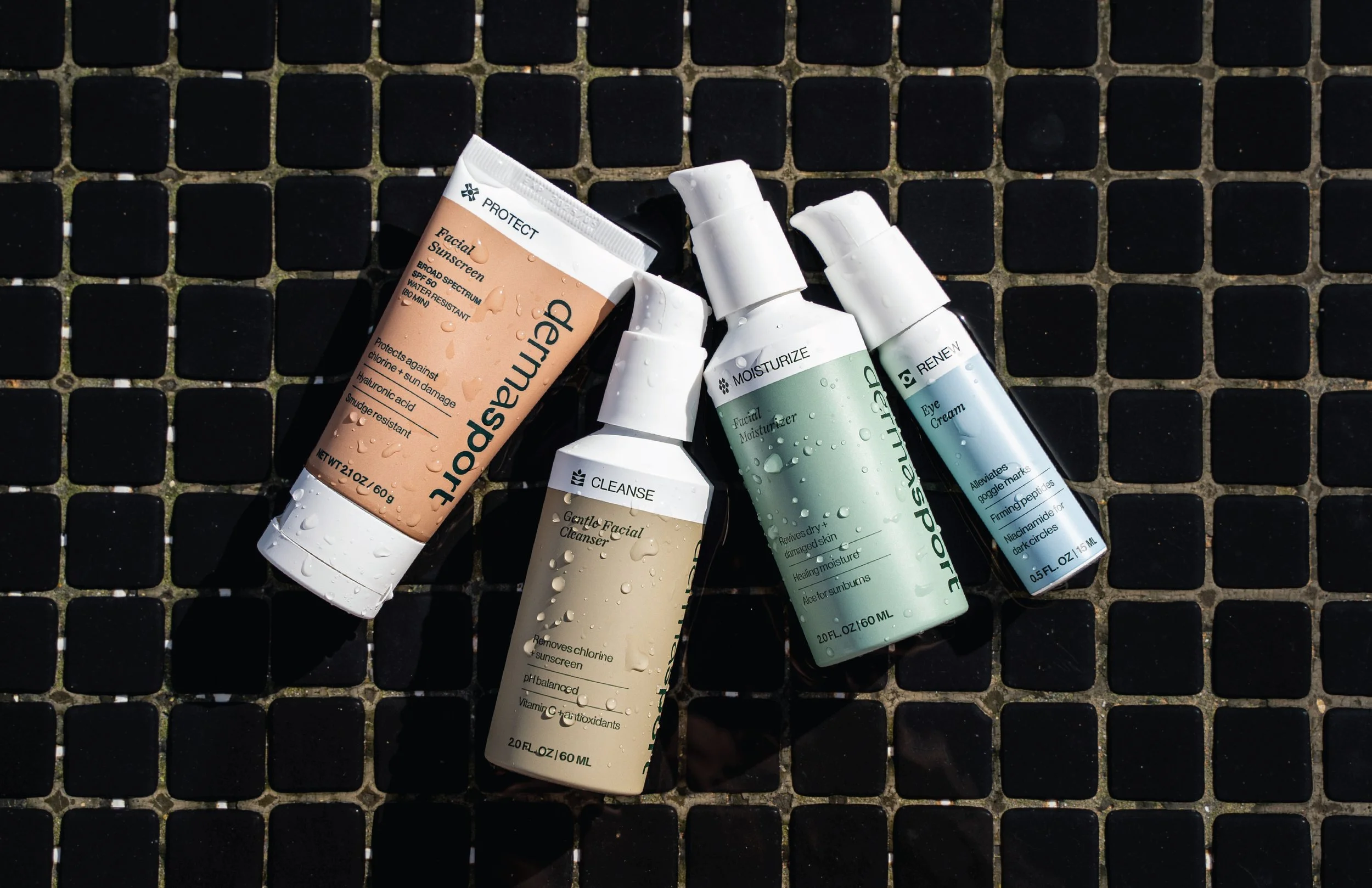 Several skincare products including facial moisturizer, eye cream, and facial cleanser, lying on a black tiled surface with water droplets on the bottles.