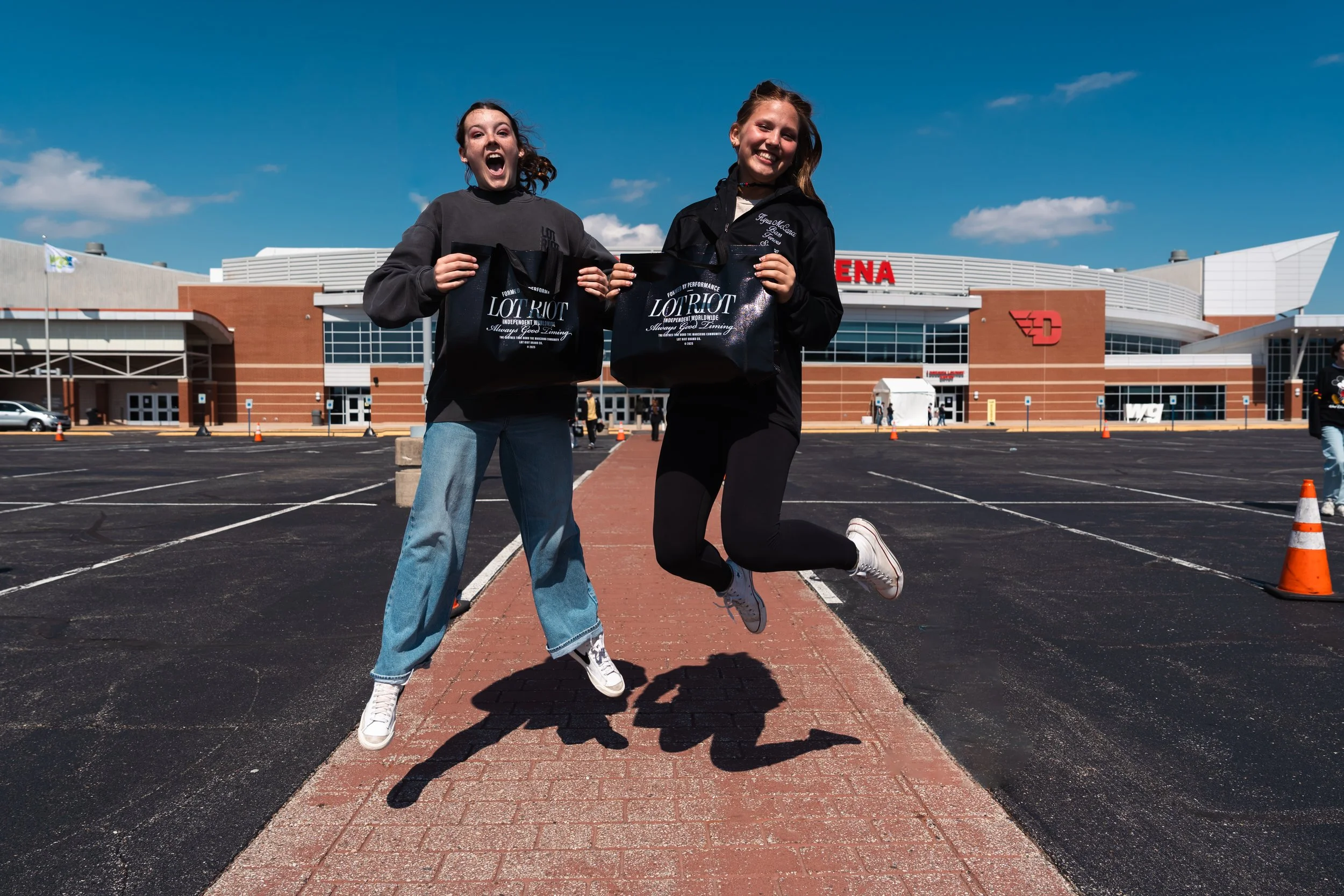 Two young women standing outside the University of Dayton for WGI Finals on a sunny day, holding black shopping bags and jumping for joy.