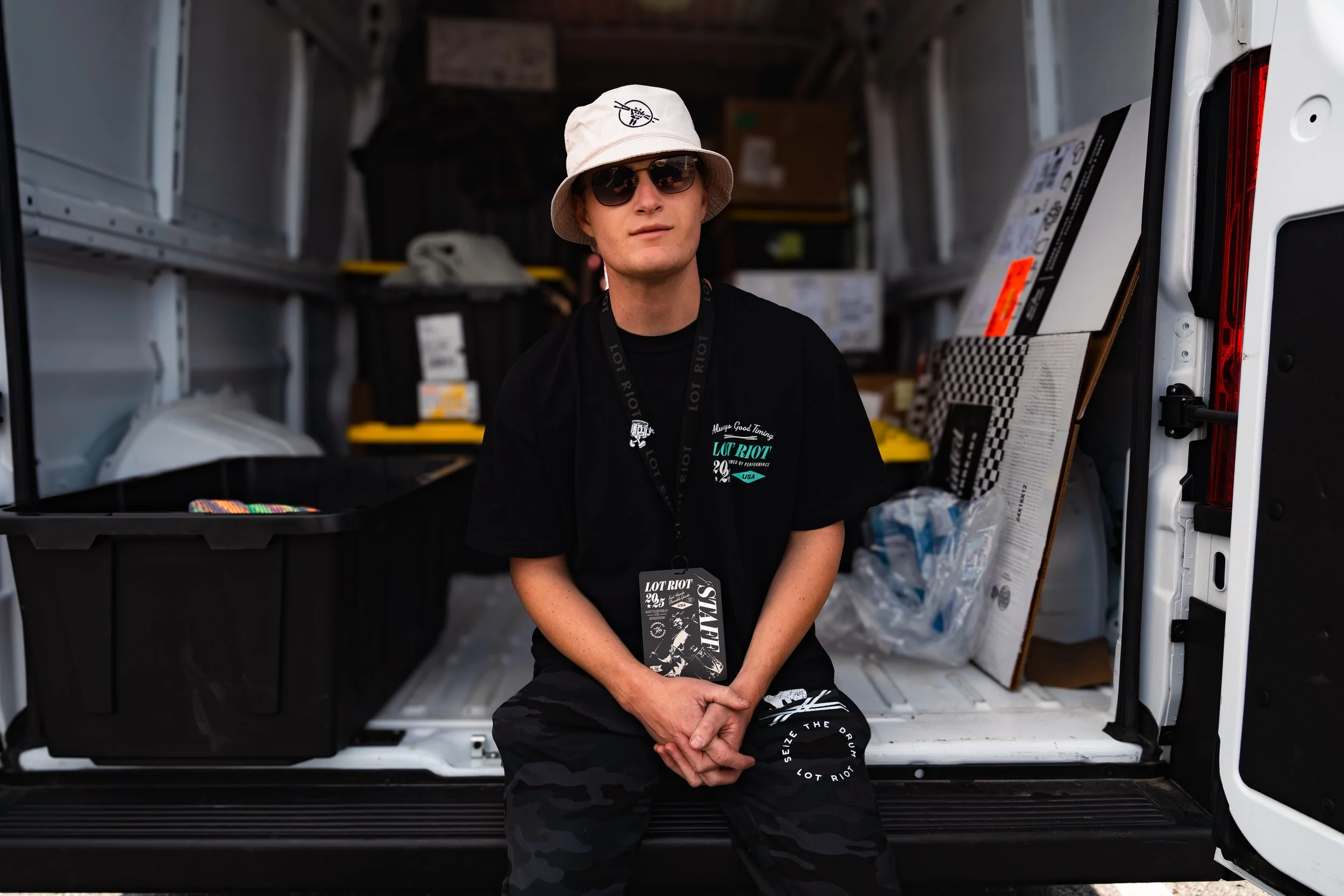 Young man sitting in the open back of a van, wearing sunglasses, a beige bucket hat with a black logo, and a black T-shirt with graphics and text, surrounded by boxes and supplies.