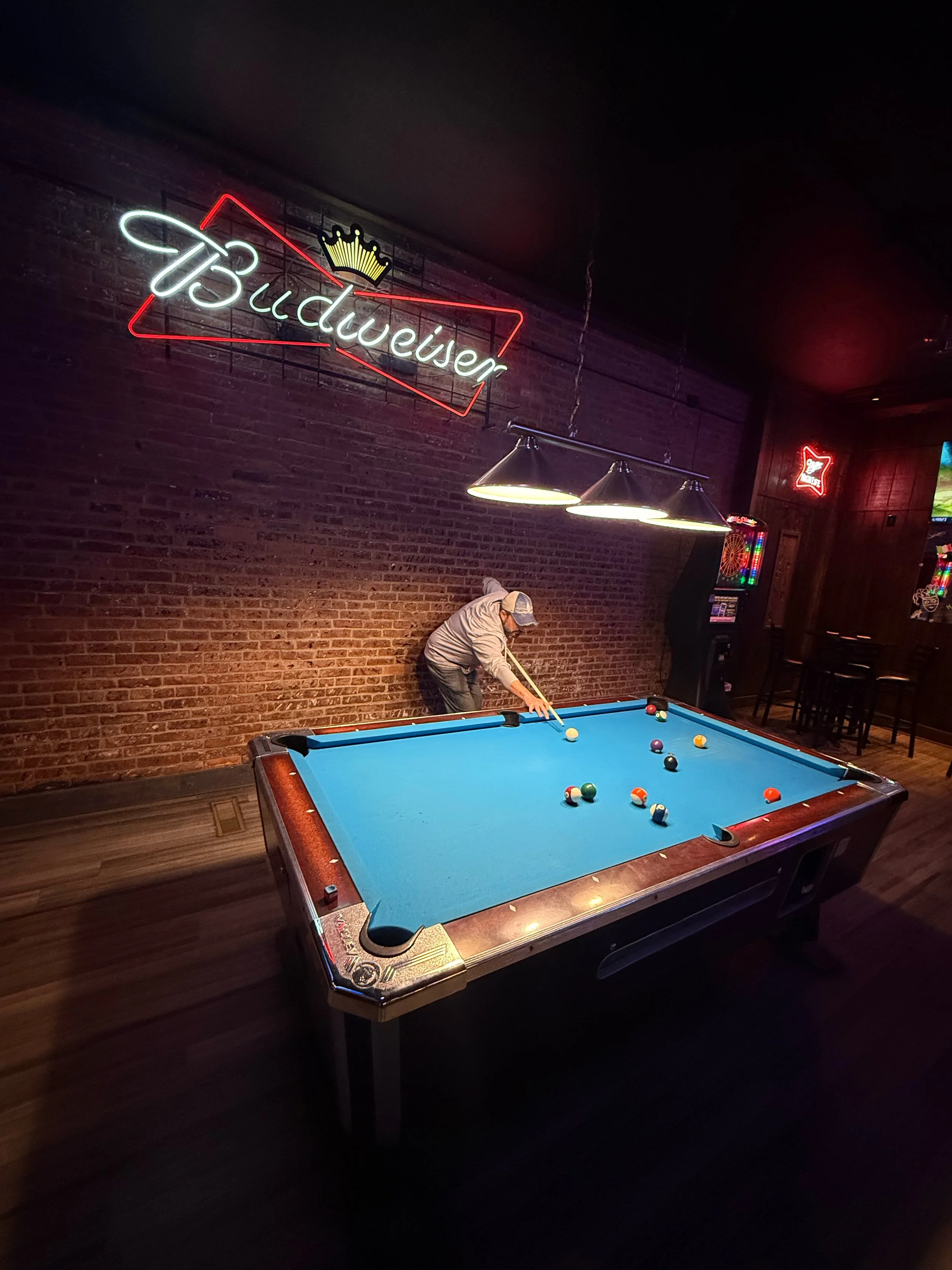 Indoor scene with a blue pool table, a person playing pool, neon signs reading 'Budweiser' and a crown, and bar seating.