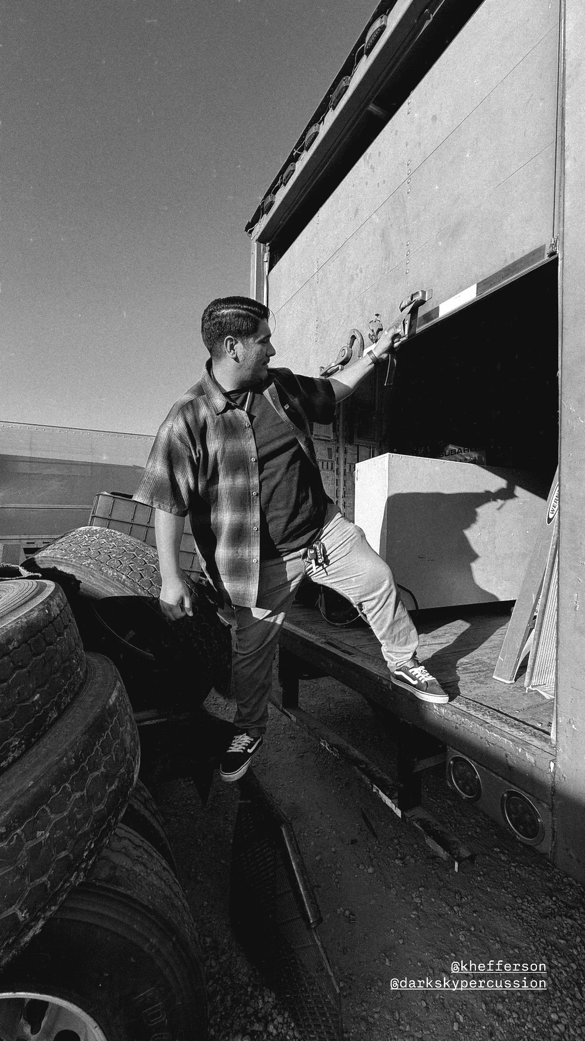 A man loading drums into a truck at a tire yard. The scene is outdoor, sunny, and black-and-white. The man is wearing a short-sleeve checkered shirt and sneakers.