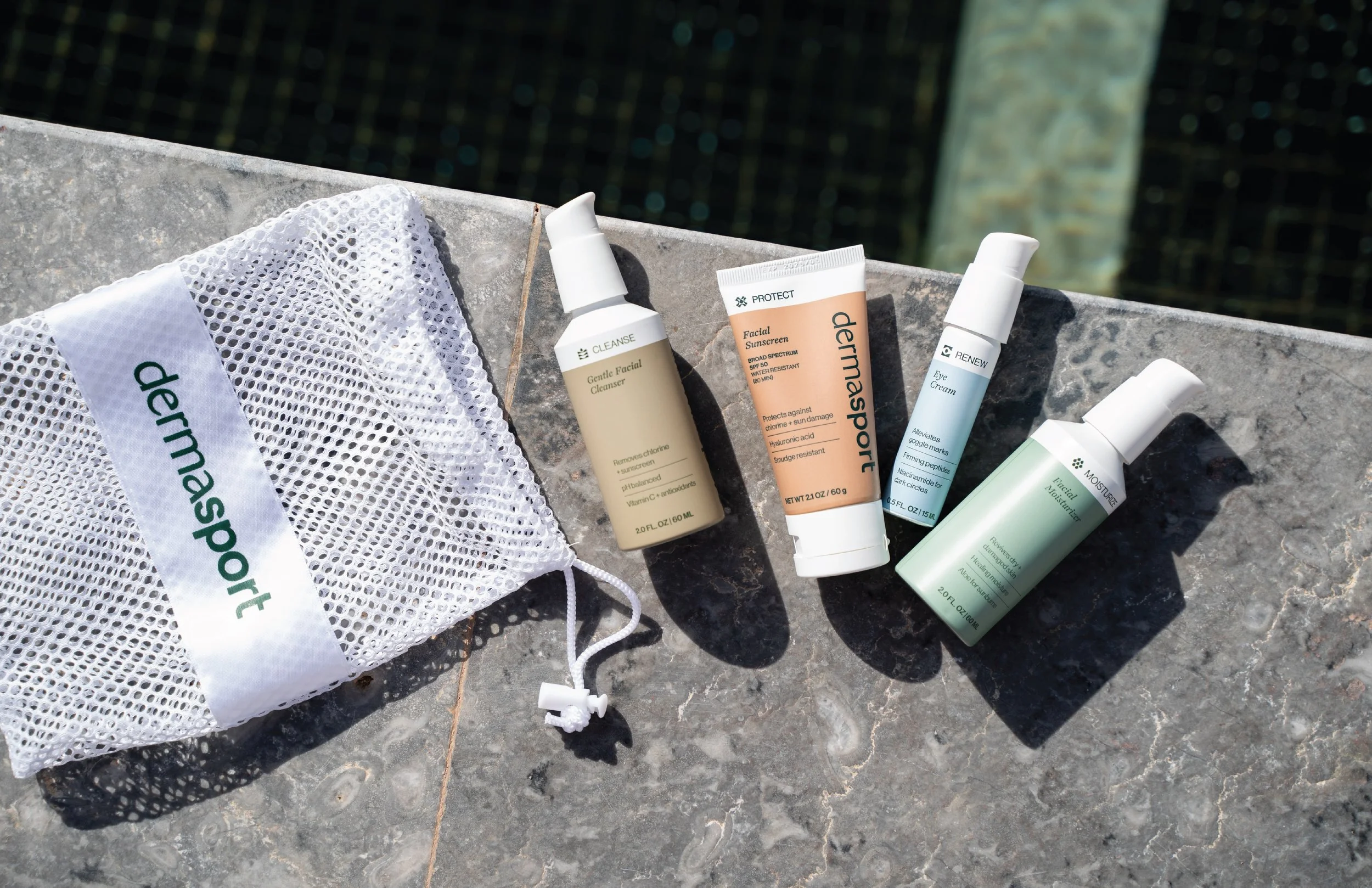 Assorted skincare and facial products, including cleansers, sunscreen, eye cream, and moisturizer, placed on a stone surface near a swimming pool, with a white mesh bag labeled 'dermask' nearby.