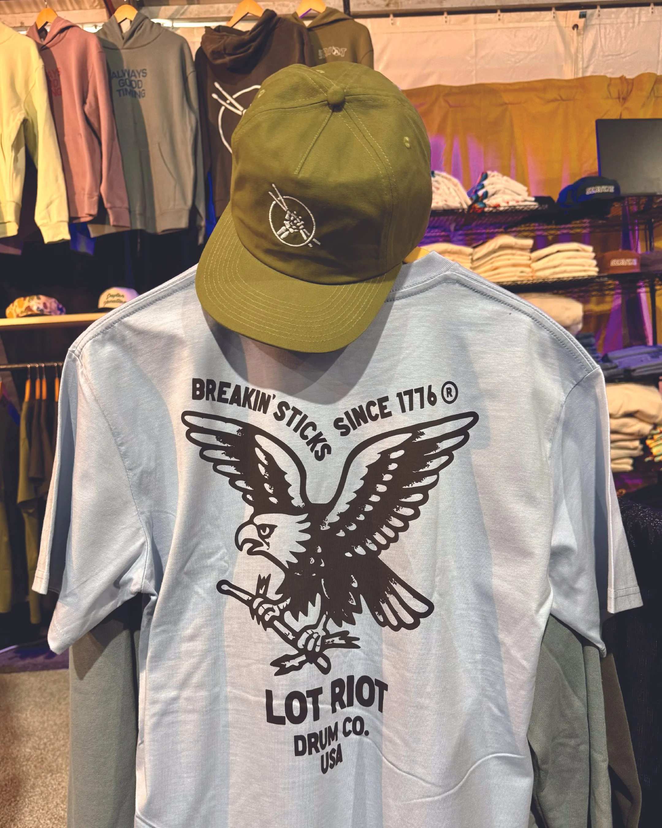 A white sweatshirt with a large black graphic of an eagle holding a drumstick in its talon, and a smaller drumstick in its beak. The sweatshirt has black text that reads 'BREAKIN' STICK SINCE 1776 LOT RIOT DRUM CO. USA'. A green cap with a white embr