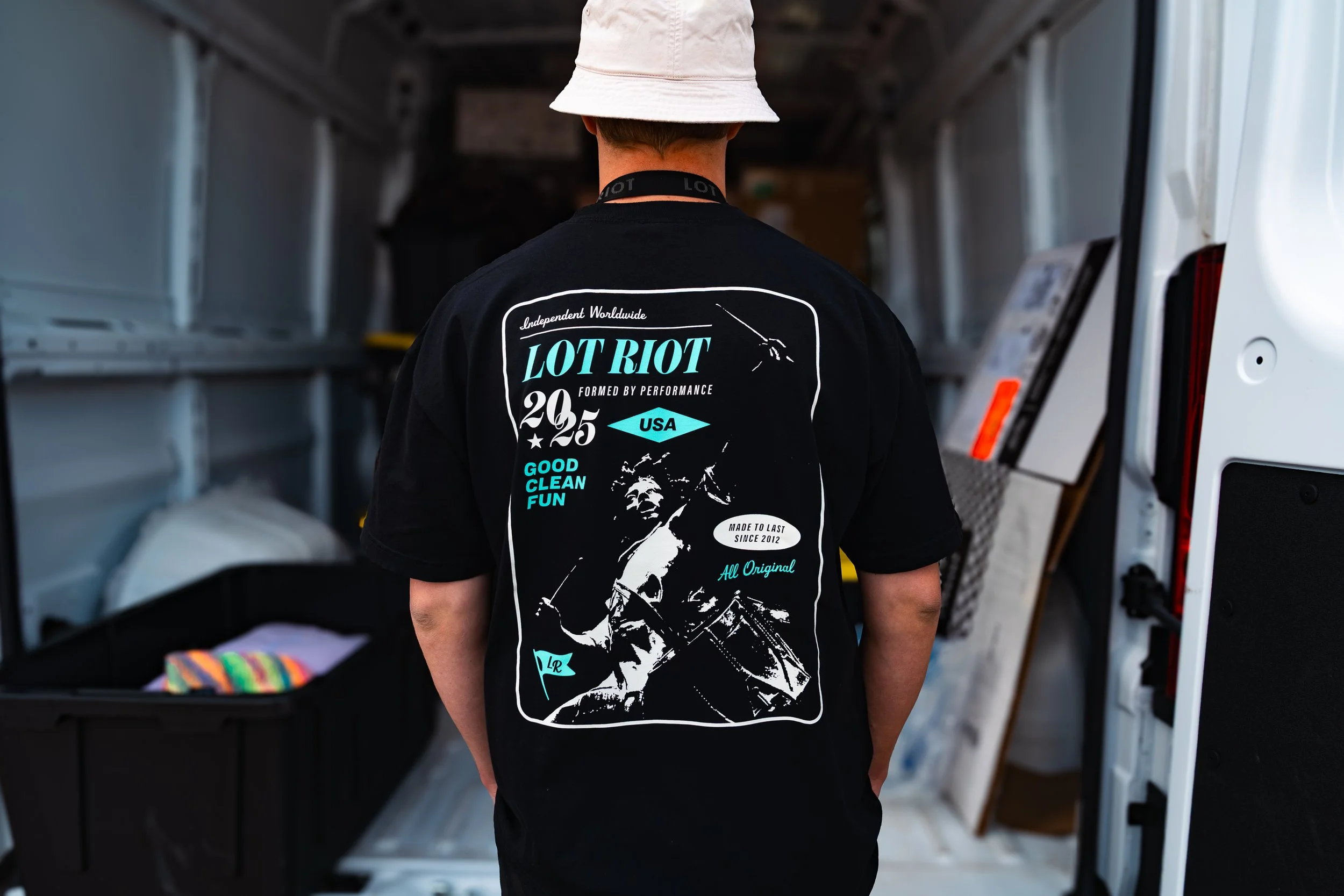 Back of a person wearing a black graphic t-shirt with text and images, standing in front of open vehicle doors with shelves and objects inside.
