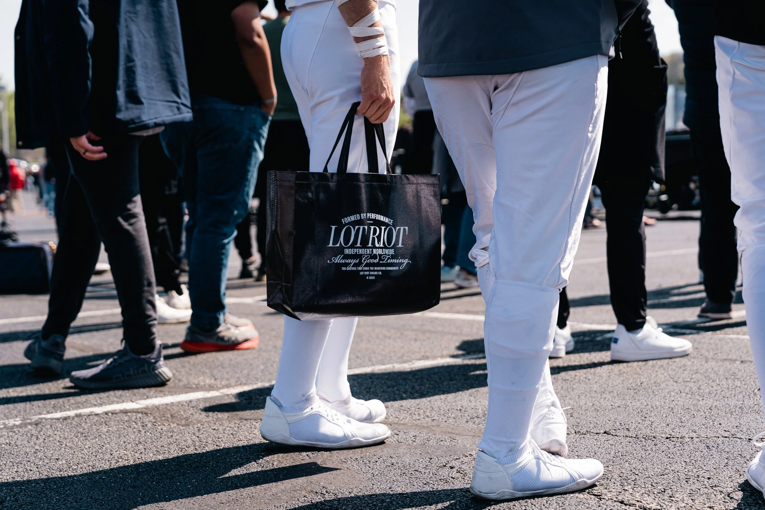 People standing in the WGI Finals lot, with one person holding a black tote bag that reads 'LOTRIOT' in white lettering, dressed in white sneakers and pants.