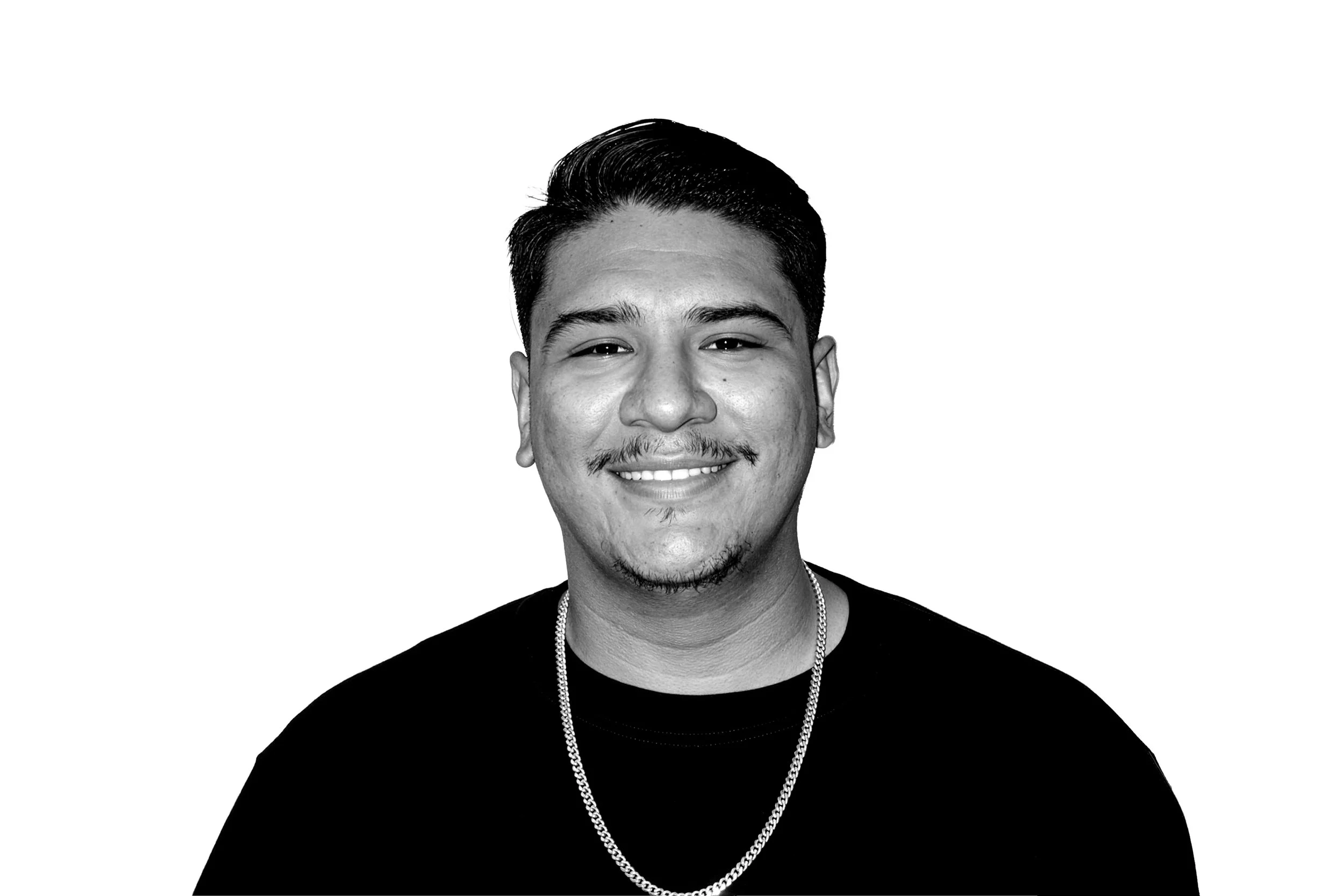 Black and white portrait of a smiling man with styled hair, mustache, and goatee, wearing a black shirt and a chain necklace.