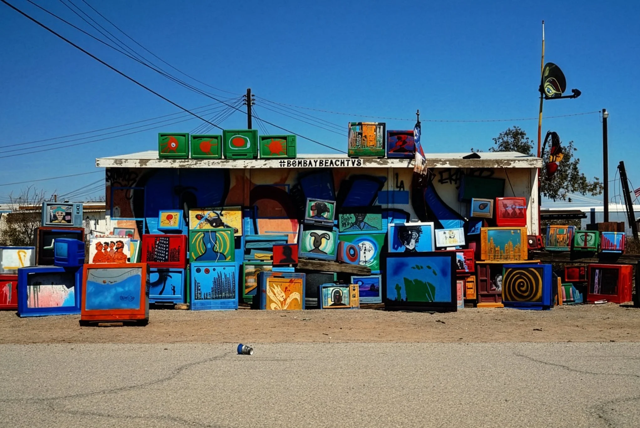 Outdoor art market with colorful paintings and framed artworks displayed on tables and a building. Clear blue sky above with power lines in the background.