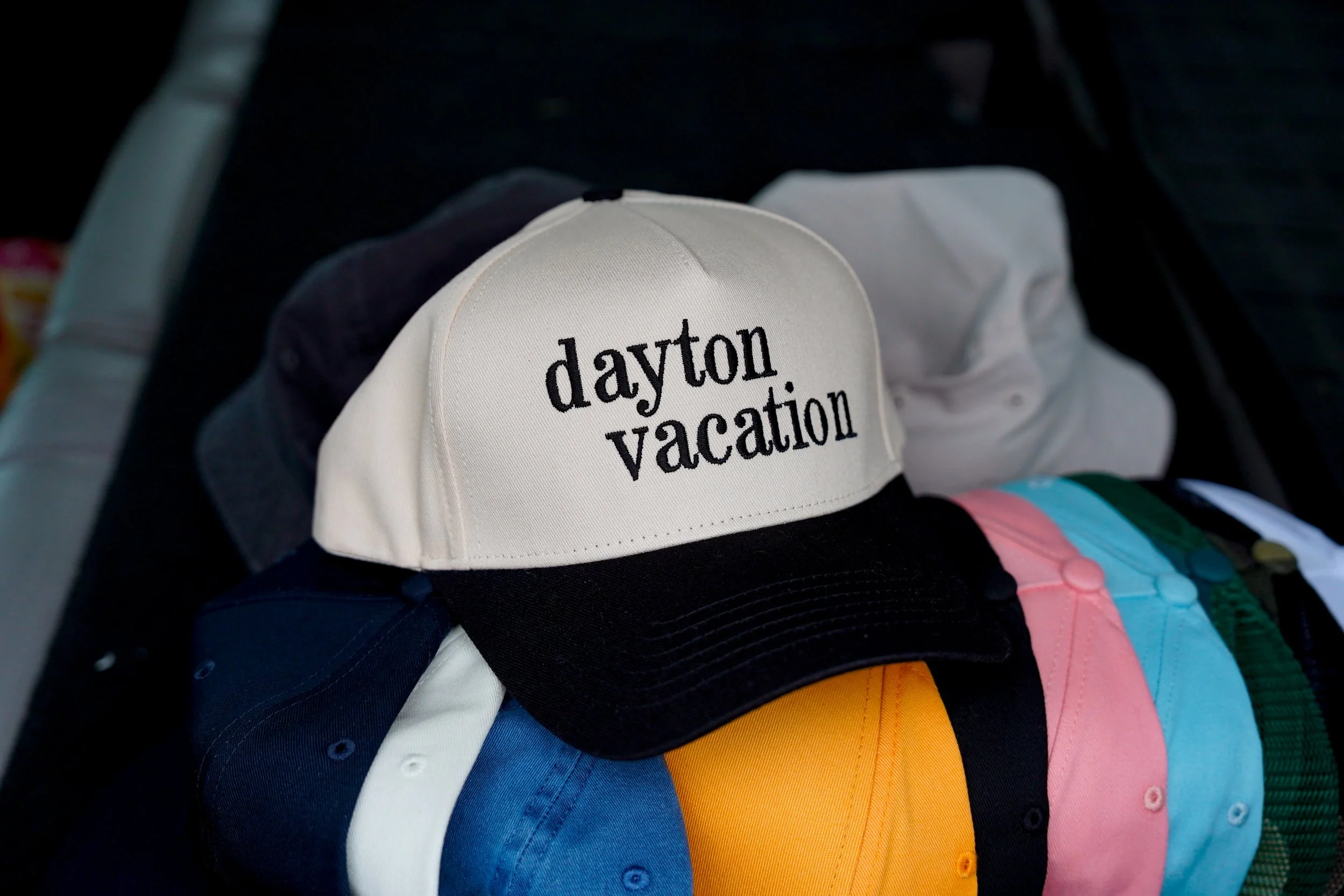 A display of caps arranged in rainbow order on a rack. A white cap with black text saying 'dayton vacation' is prominent in the front.