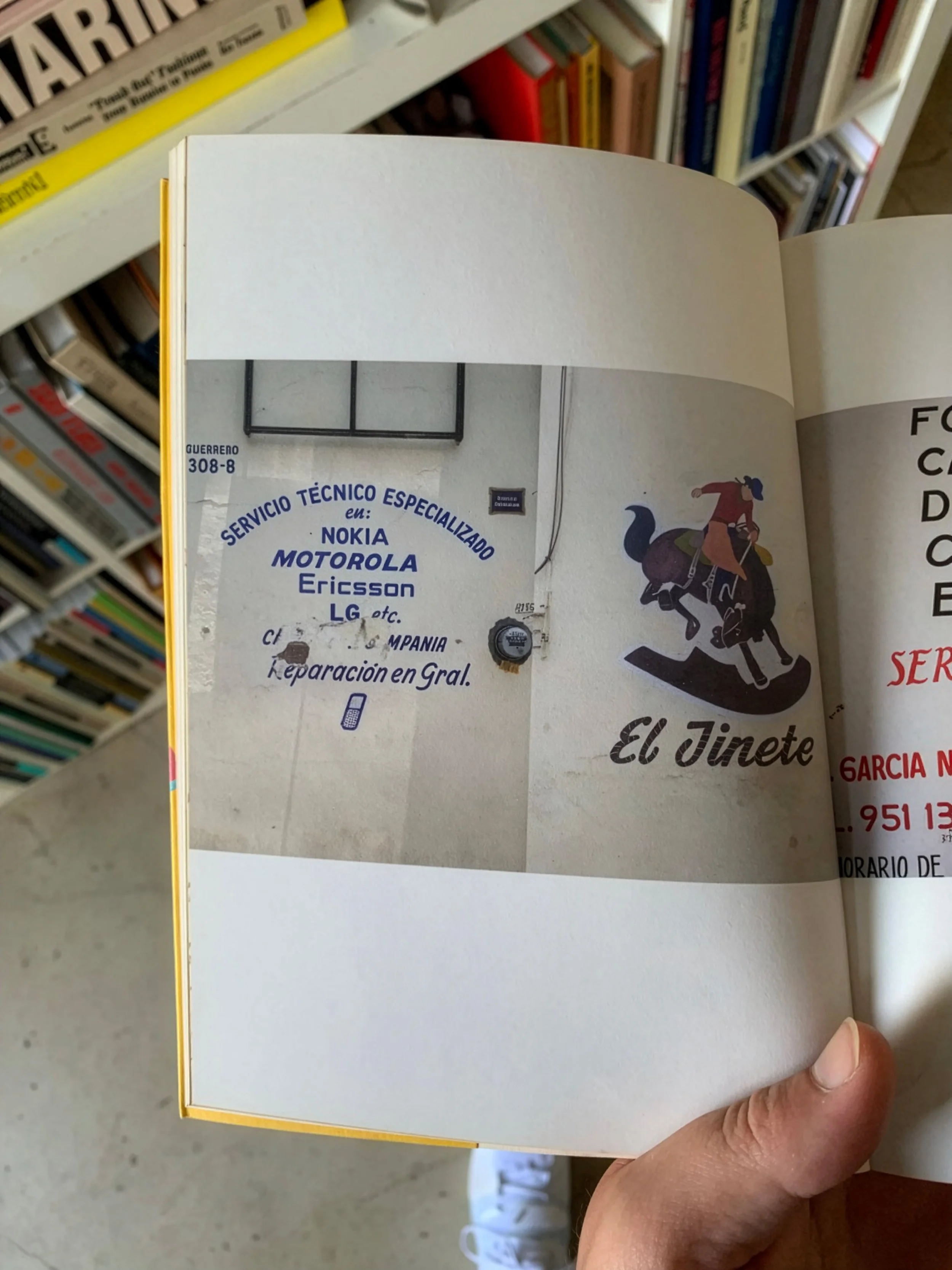 A hand holding a book open to a page with a partial image and Spanish text about technical service for mobile phone brands, with bookshelves in the background.