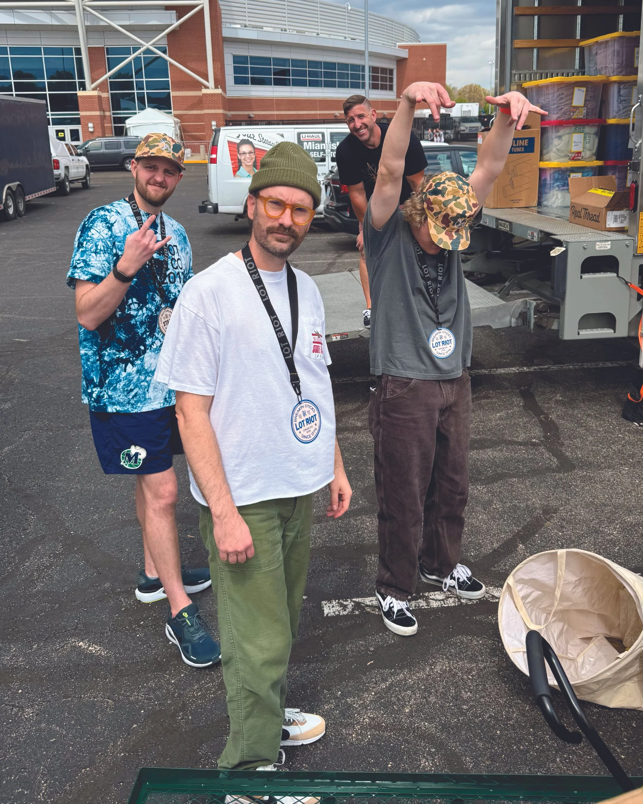 Four men wearing lanyards, with a U-Haul truck and parked cars in the background. One man is making a peace sign, another is wearing orange glasses and a green beanie, a third is lifting his arm above his head in a gesture, and the fourth is smiling 