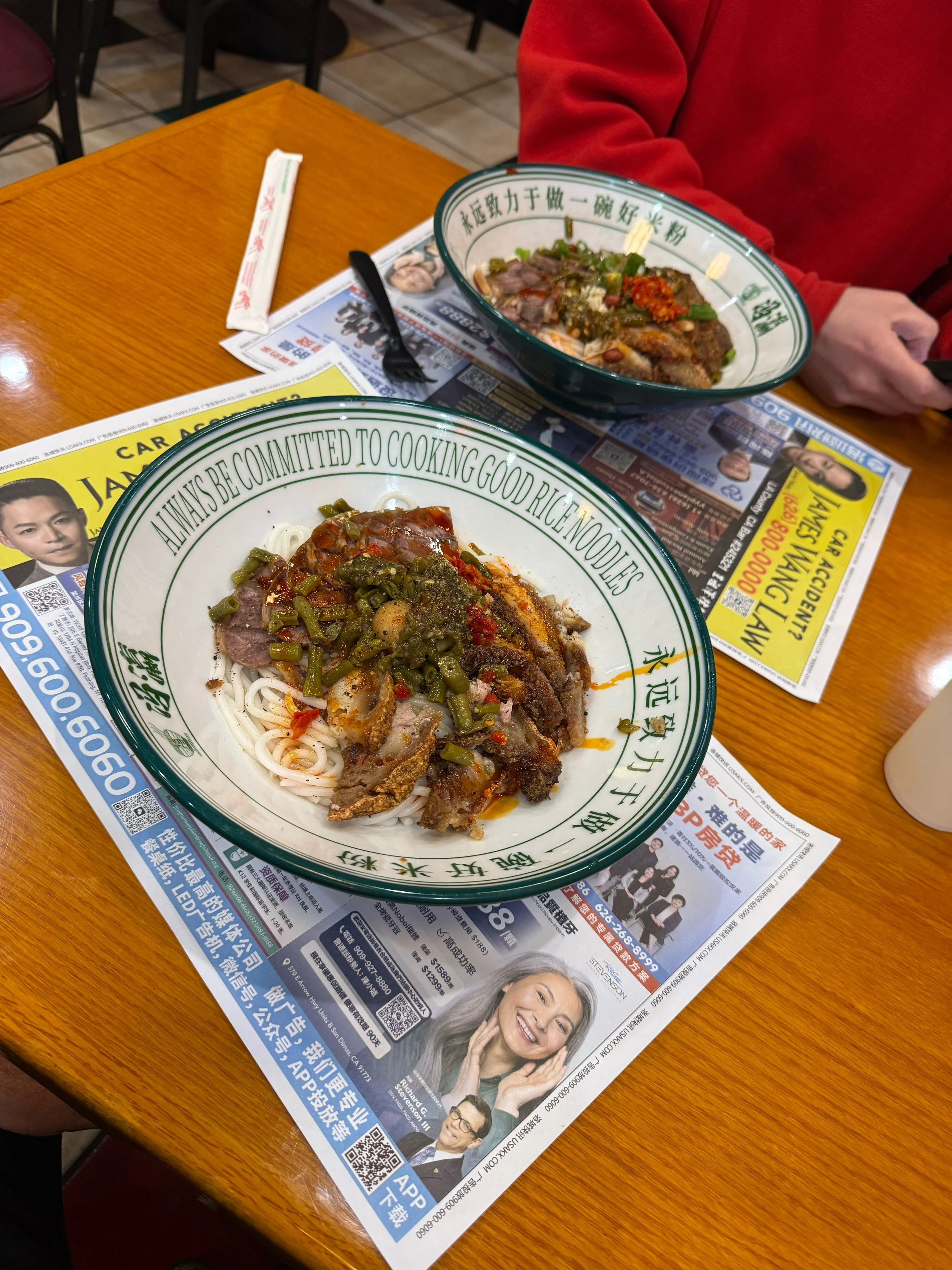 Two bowls of Asian noodle soup with sliced meat, vegetables, and chili, placed on top of classified advertisements with Chinese text on a wooden table, next to a person in a red sleeve.