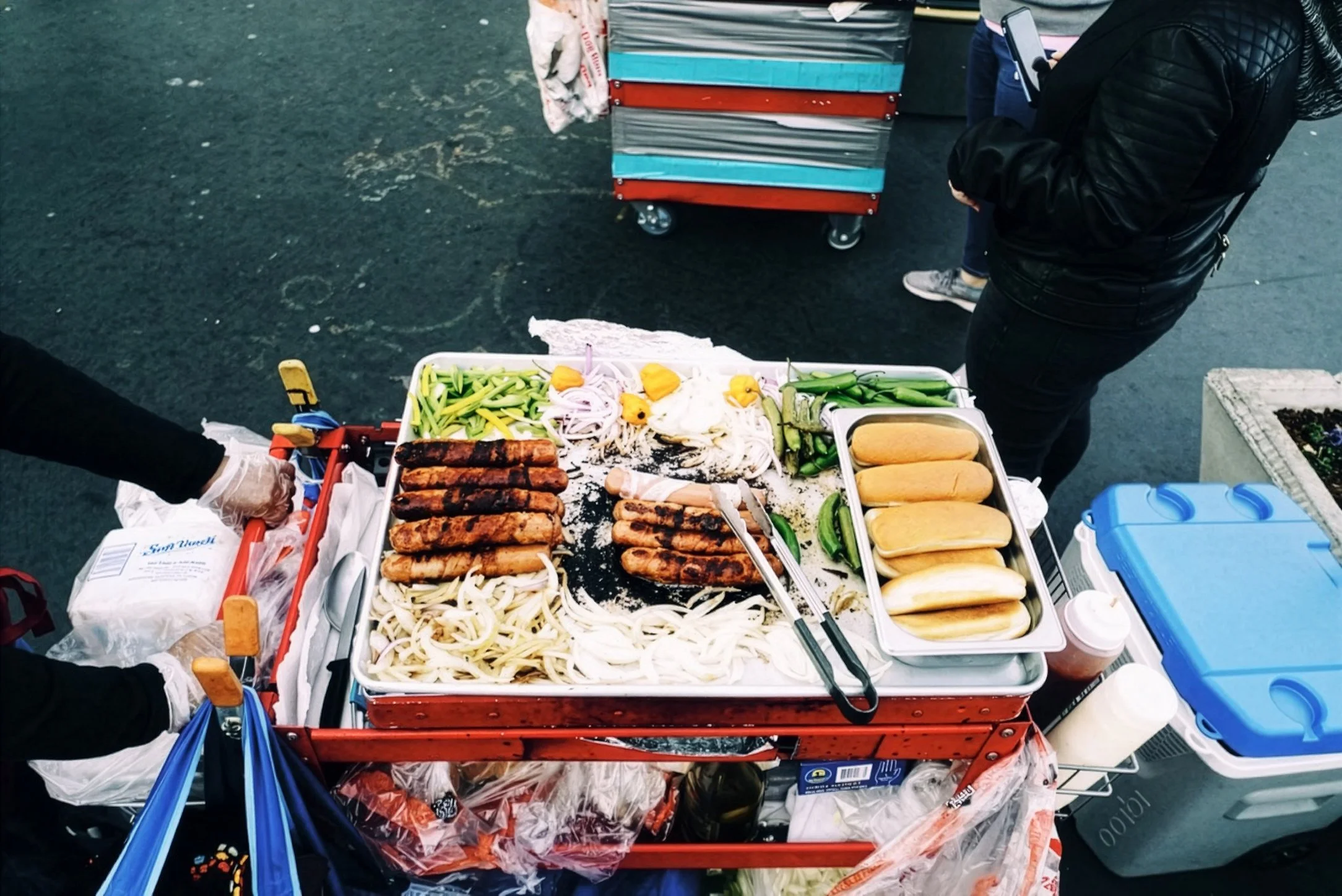 A street food cart with grilled sausages, sliced onions, and vegetables, with a person preparing food and another person standing nearby, holding a smartphone.