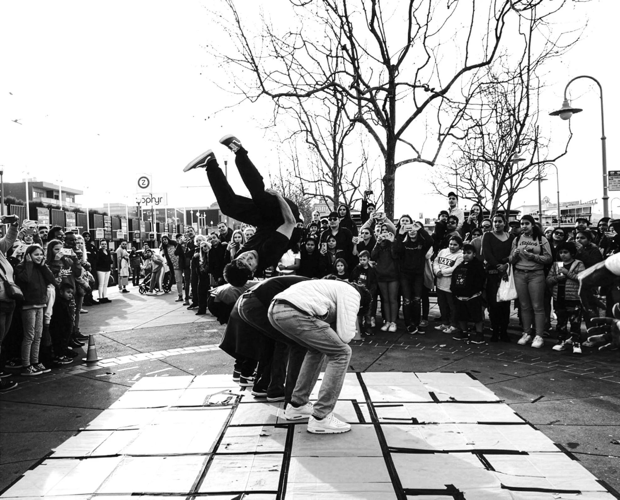 Black and white photo of a street performer doing a headstand on a platform in front of a crowd, with leafless trees and street lamps in the background.