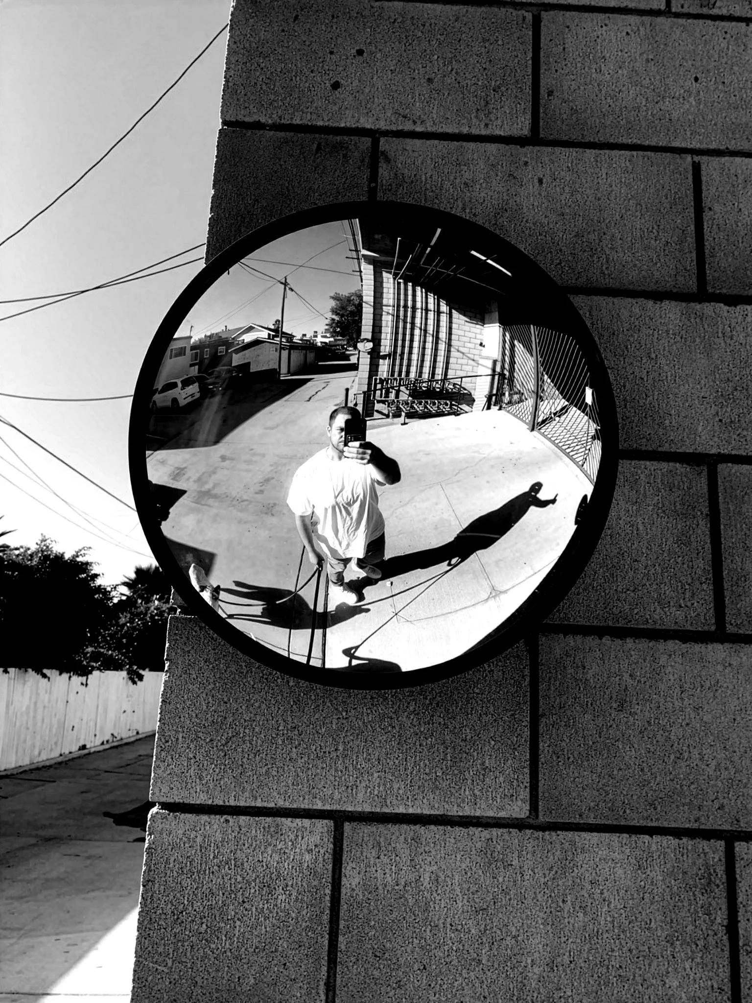 A circular convex mirror attached to a textured brick wall reflects a man taking a selfie outside on a sunny day, with residential buildings, utility poles, wires, and a fence visible in the background.
