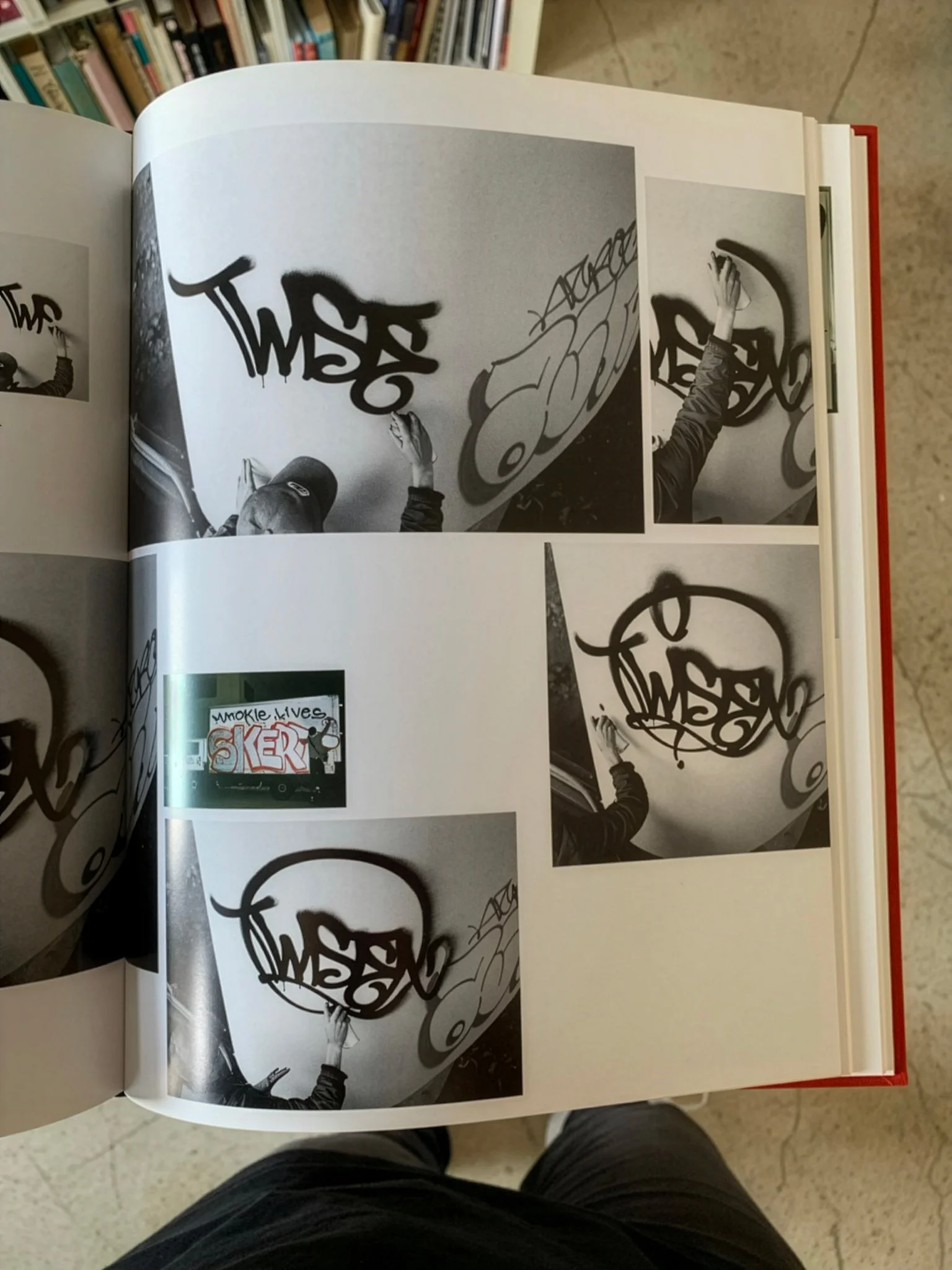 Black and white photograph of a person spray painting graffiti on a wall, with the person's hand reaching out and holding a spray can. The graffiti appears to spell out 'TOOS' or a similar word.
