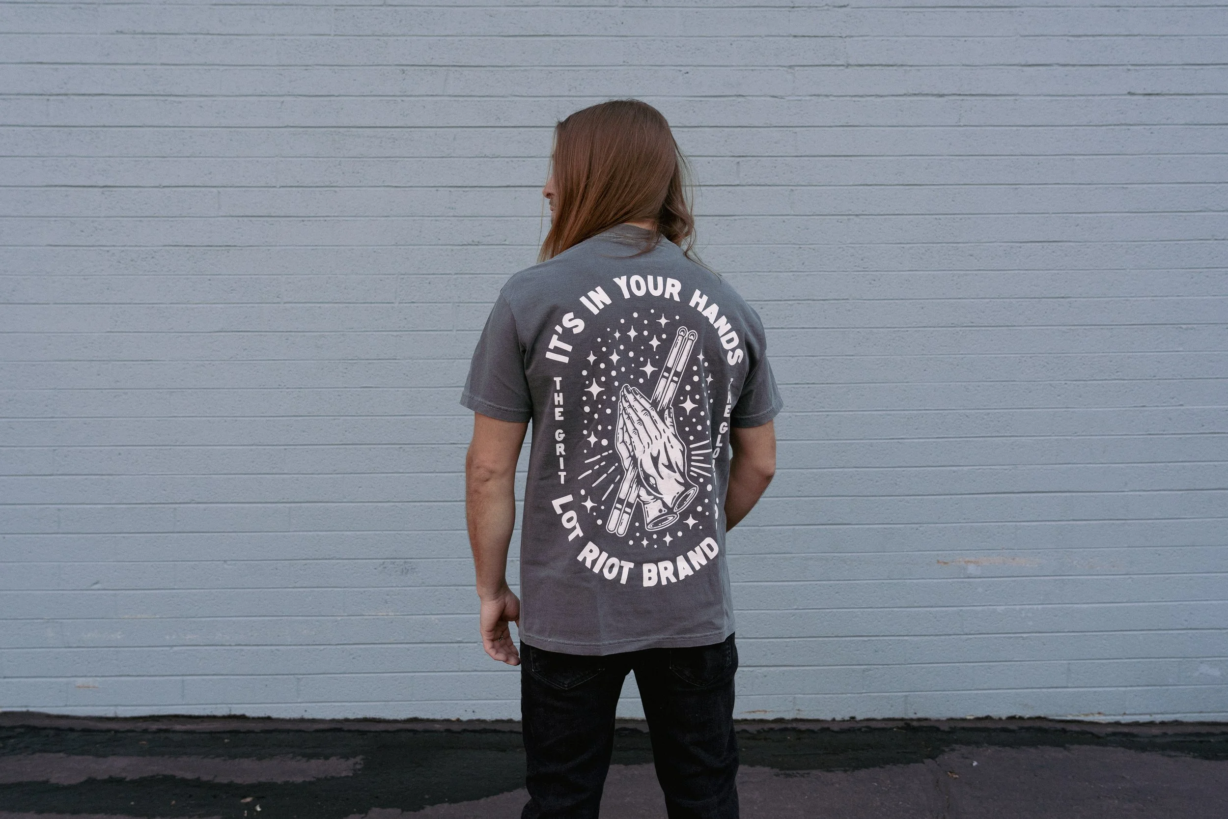 Person with long hair wearing a gray t-shirt standing against a light gray brick wall, facing away from the camera at WGI Finals in Dayton, Ohio.