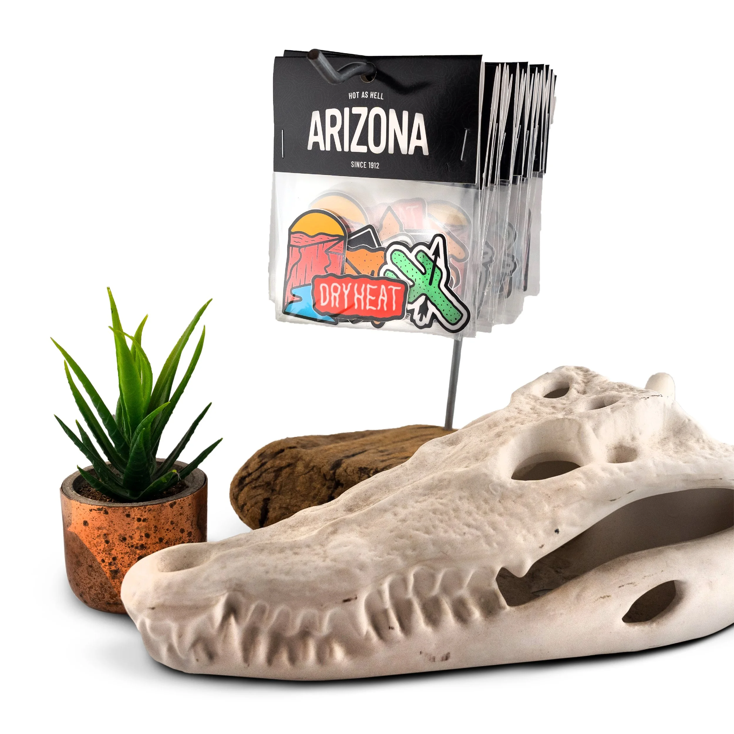 A cactus plant in a small copper pot, next to a decorative perforated white stone sculpture, with a black sign featuring stickers including a cactus, a dry heat label, and desert-themed graphics.