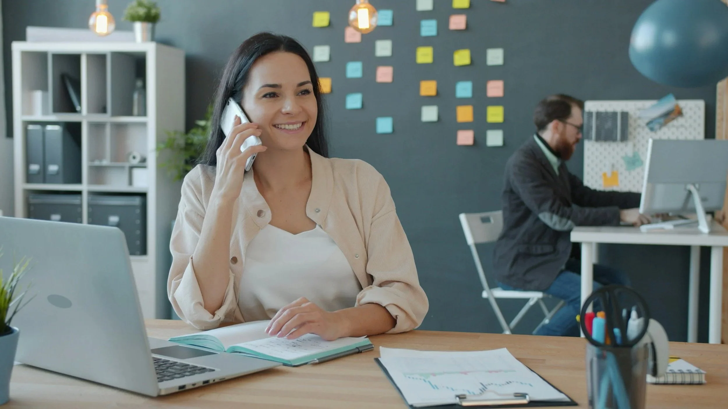 A customer support representative is smiling while talking on the phone. Her coworker is busy on a computer in the background. There are many sticky notes on the wall behind them.