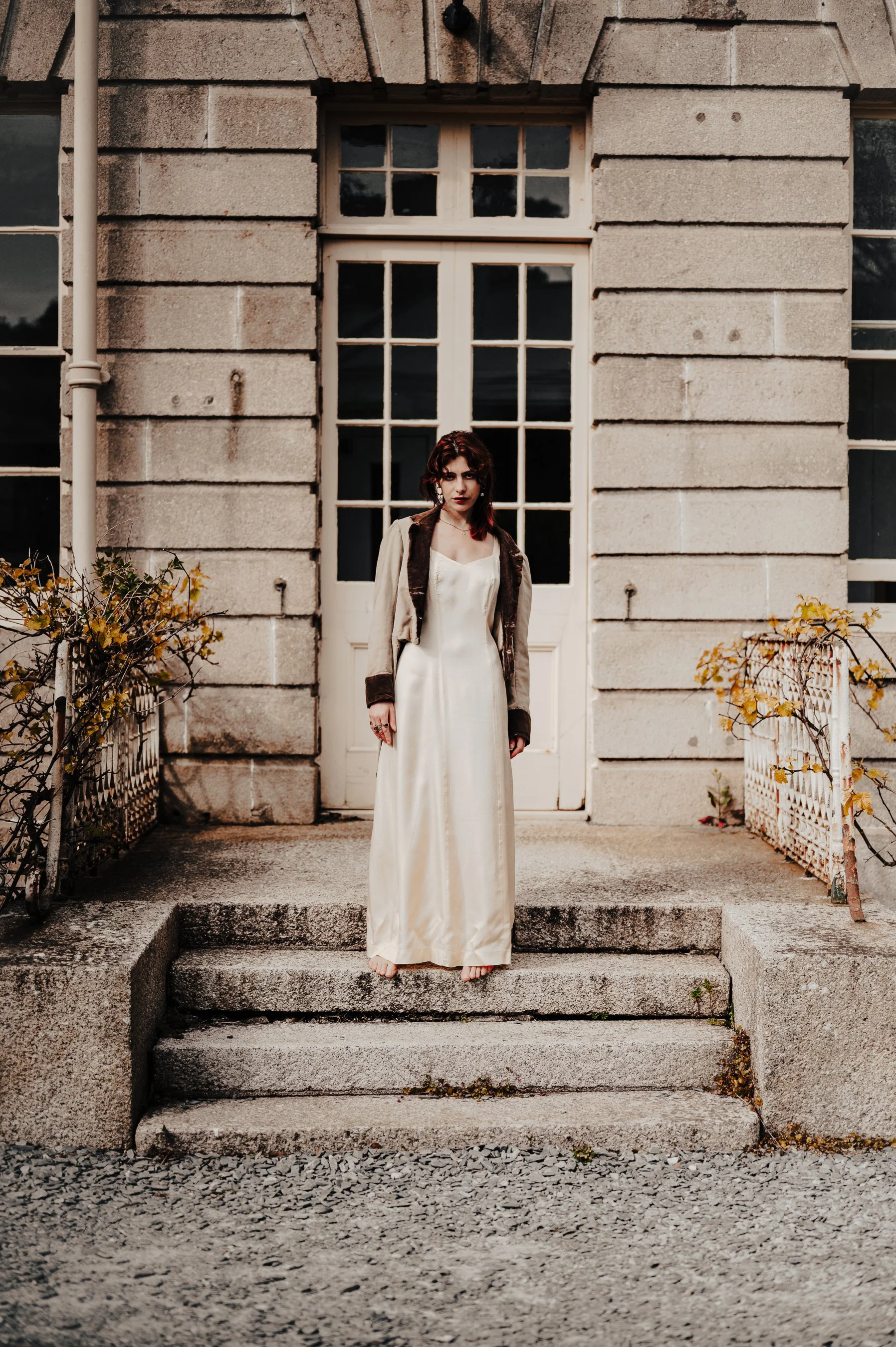 A young woman with dark red hair standing barefoot on steps in front of a stone building with large windows, wearing a long white dress and a brown jacket.
