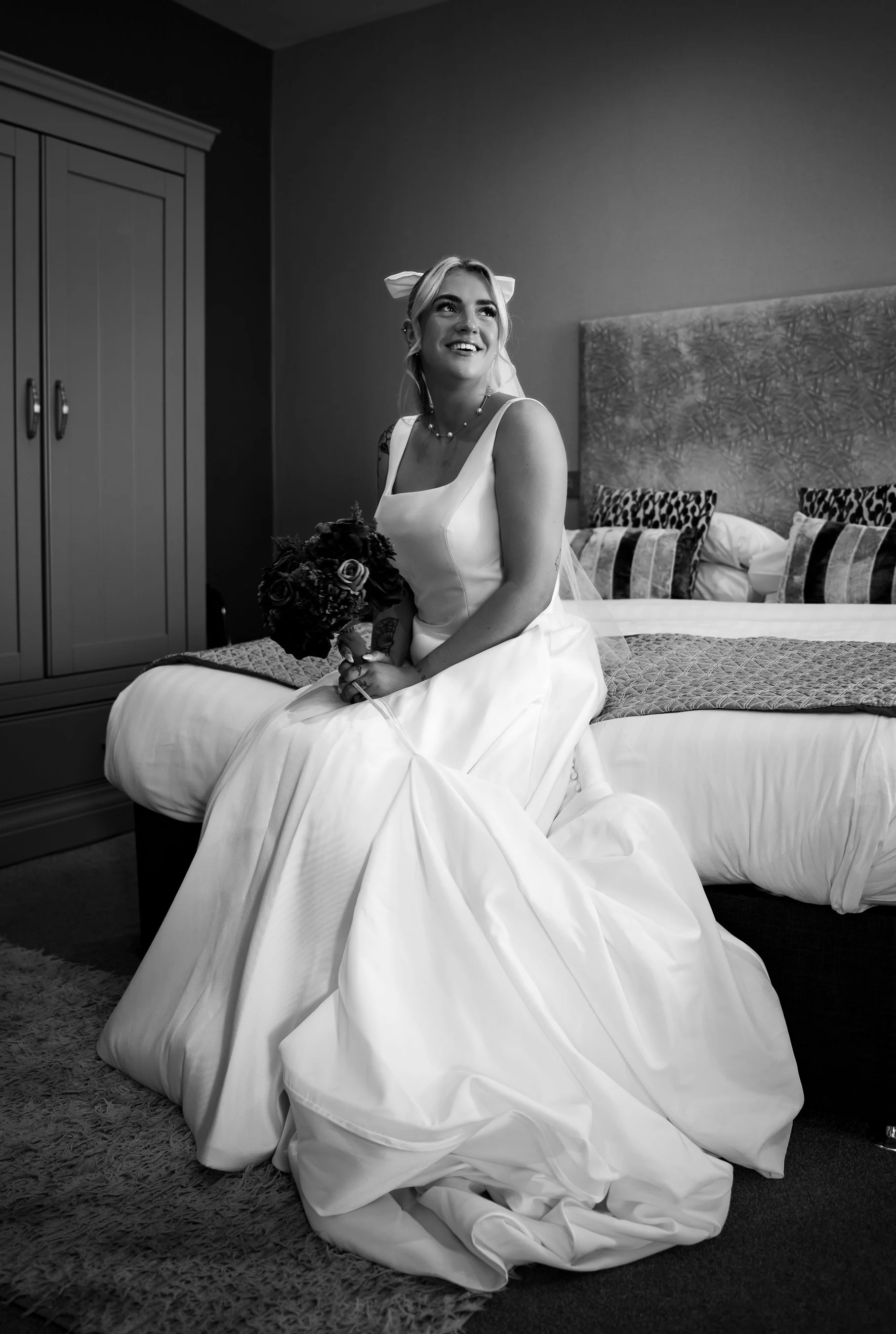 A woman in a wedding dress sitting on a bed, holding a bouquet of flowers, smiling and looking up, in a bedroom with a wardrobe and patterned pillows.