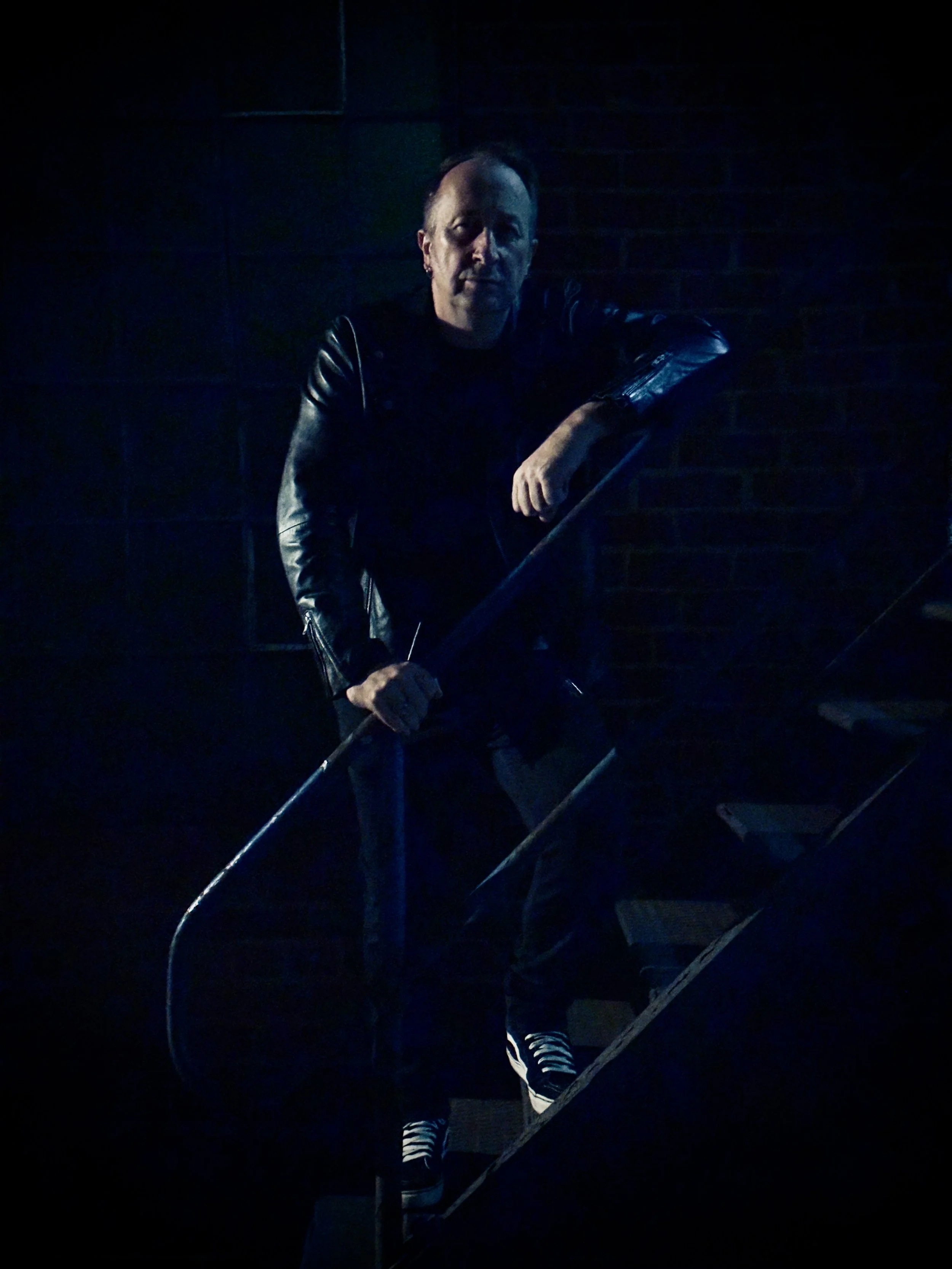 Atmospheric portrait of UK musician Paul Arden standing on a metal staircase in low blue lighting. Wearing a black leather jacket and monochrome sneakers, he leans on the railing against a brick wall backdrop, capturing his moody, urban aesthetic and