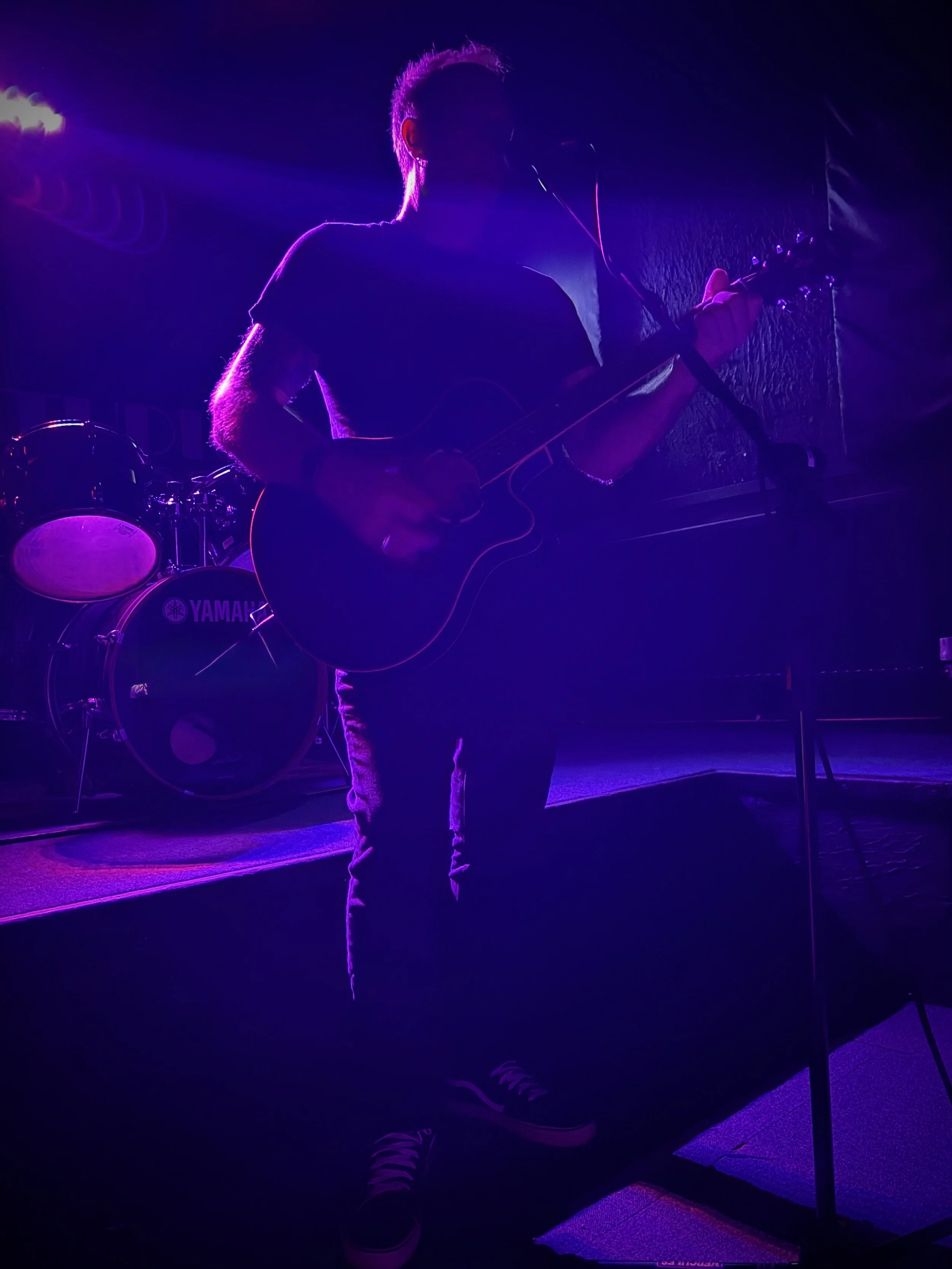 Live performance photograph of UK musician Paul Arden playing acoustic guitar under deep purple stage lighting. Captured mid‑song with microphone and drum kit in the background, the image conveys the cinematic atmosphere and intensity of his live sho