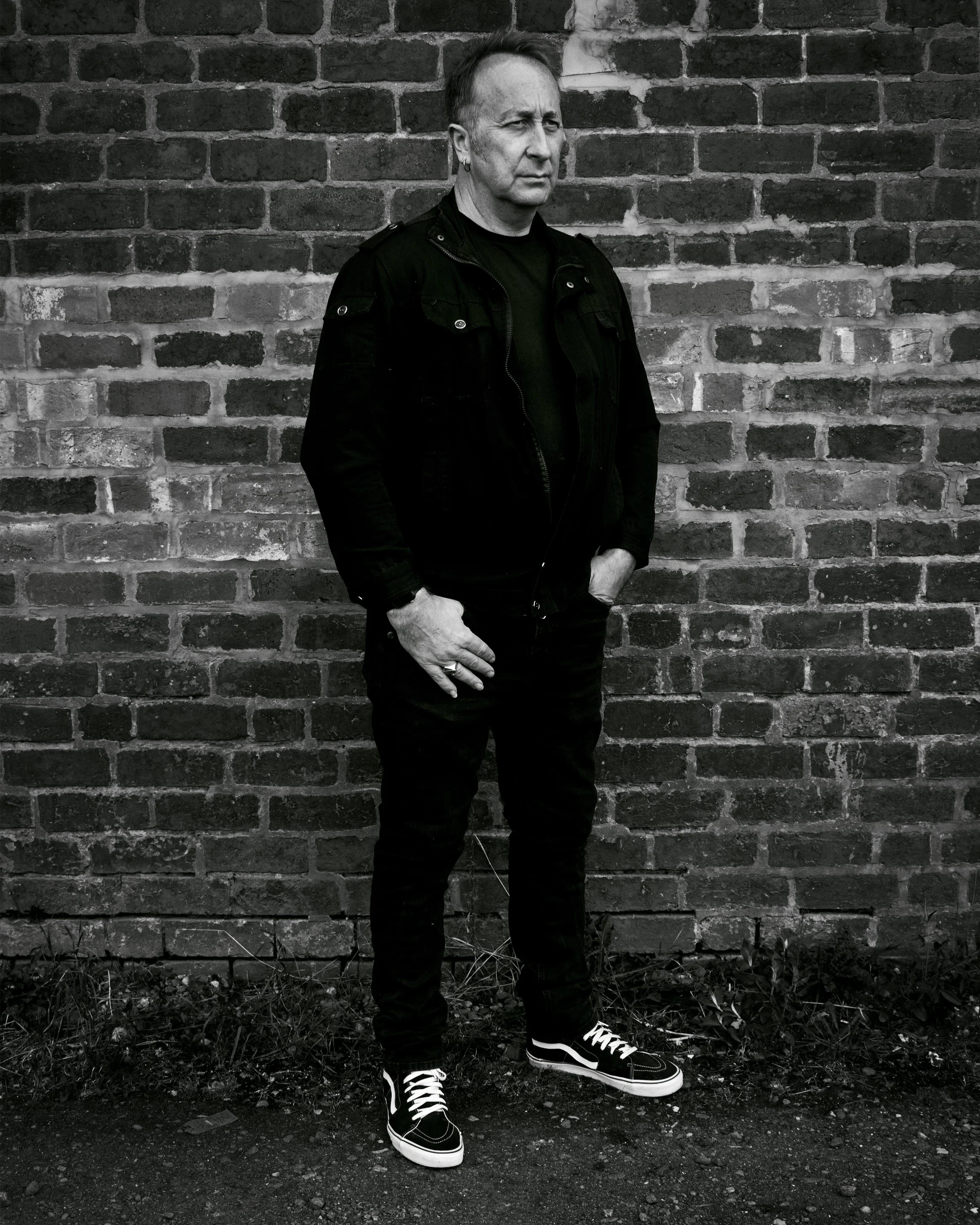 Black‑clad figure standing against a weathered brick wall, gaze turned aside — a quiet, introspective portrait with muted tones and texture.