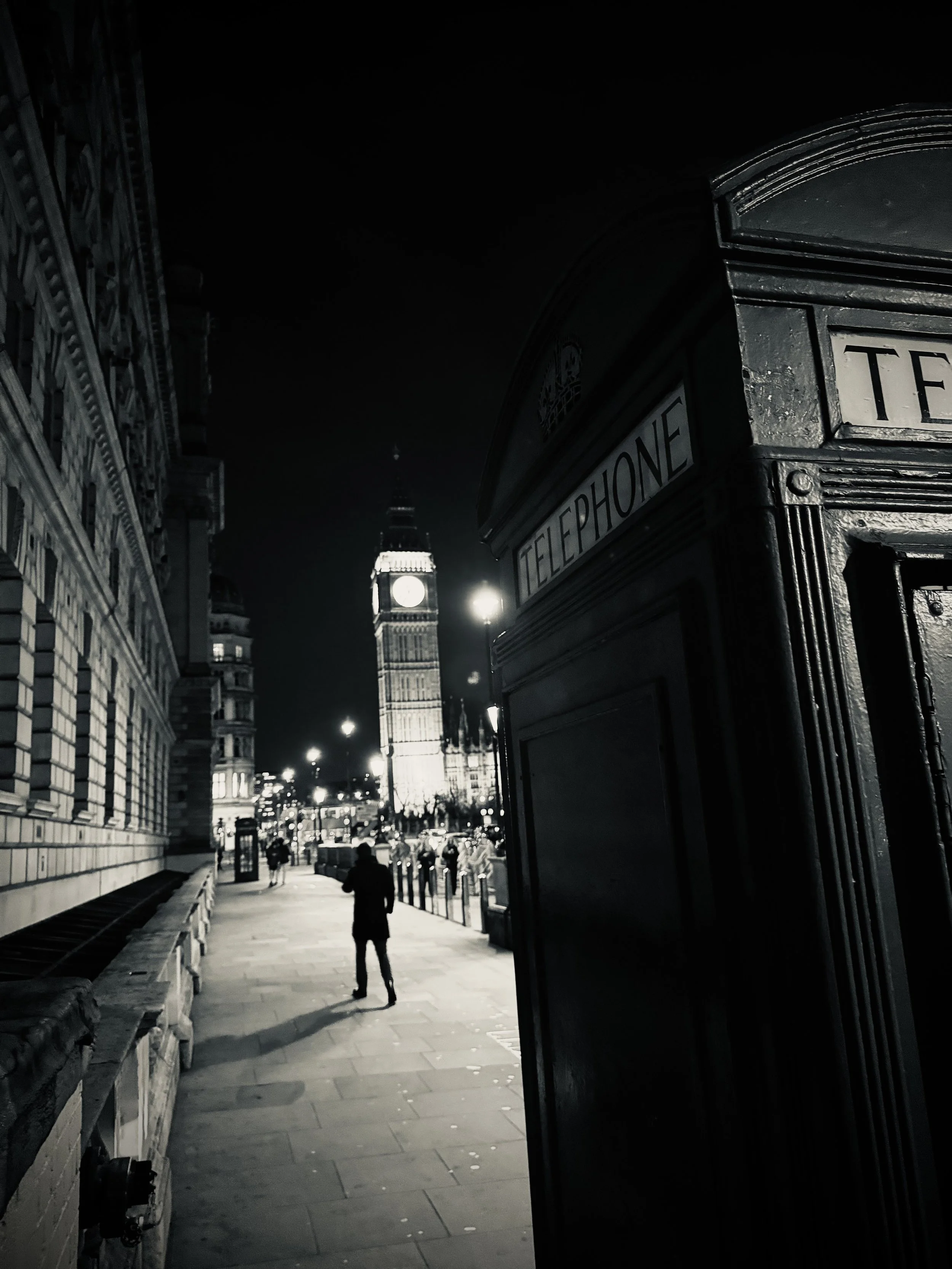 Nocturnal London street scene — a glowing telephone booth and silhouettes of passersby beneath the illuminated face of Big Ben, the city alive in shadow and light.