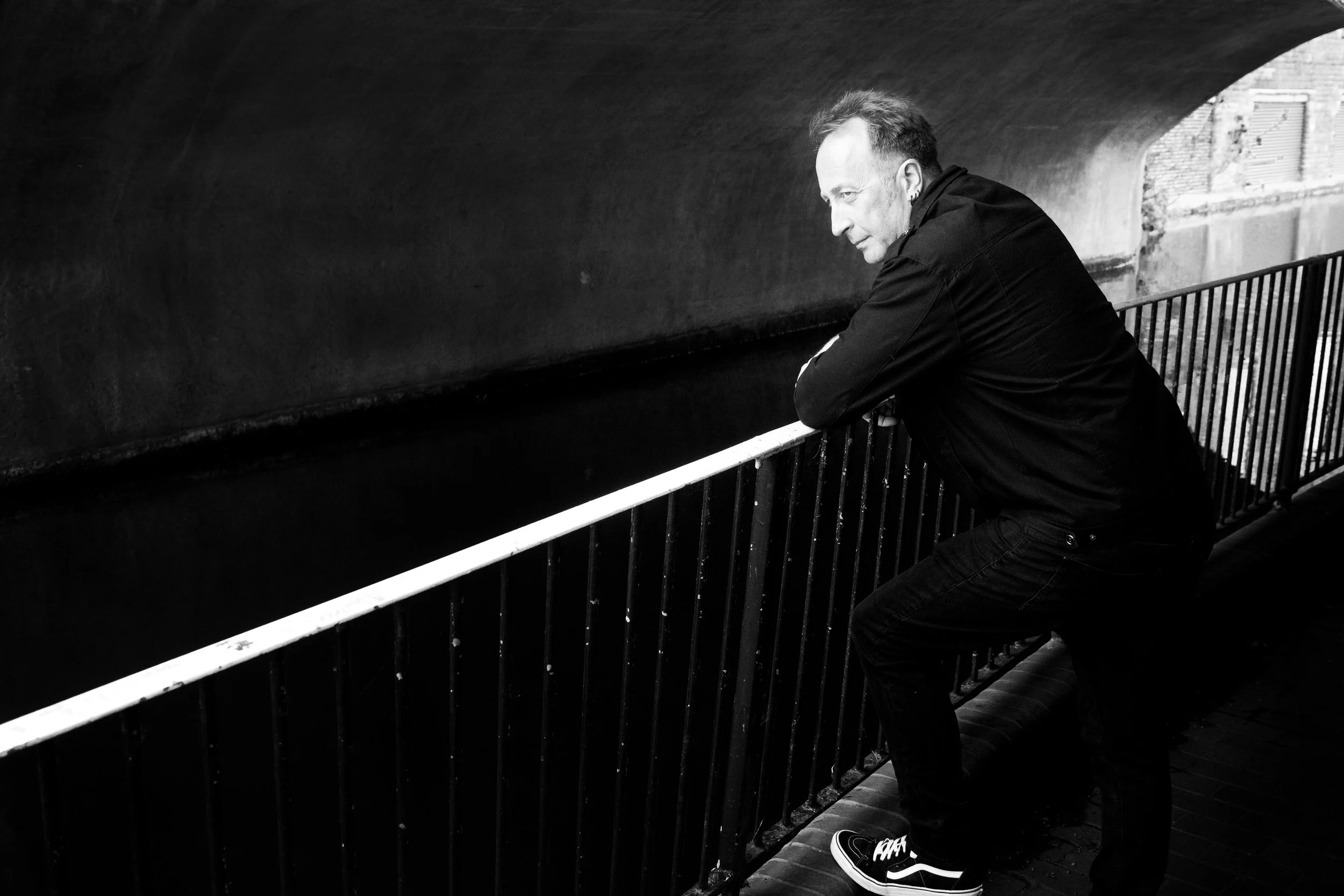 Black‑and‑white portrait of a man leaning on a railing beneath an urban bridge — moody, reflective, and cinematic.
