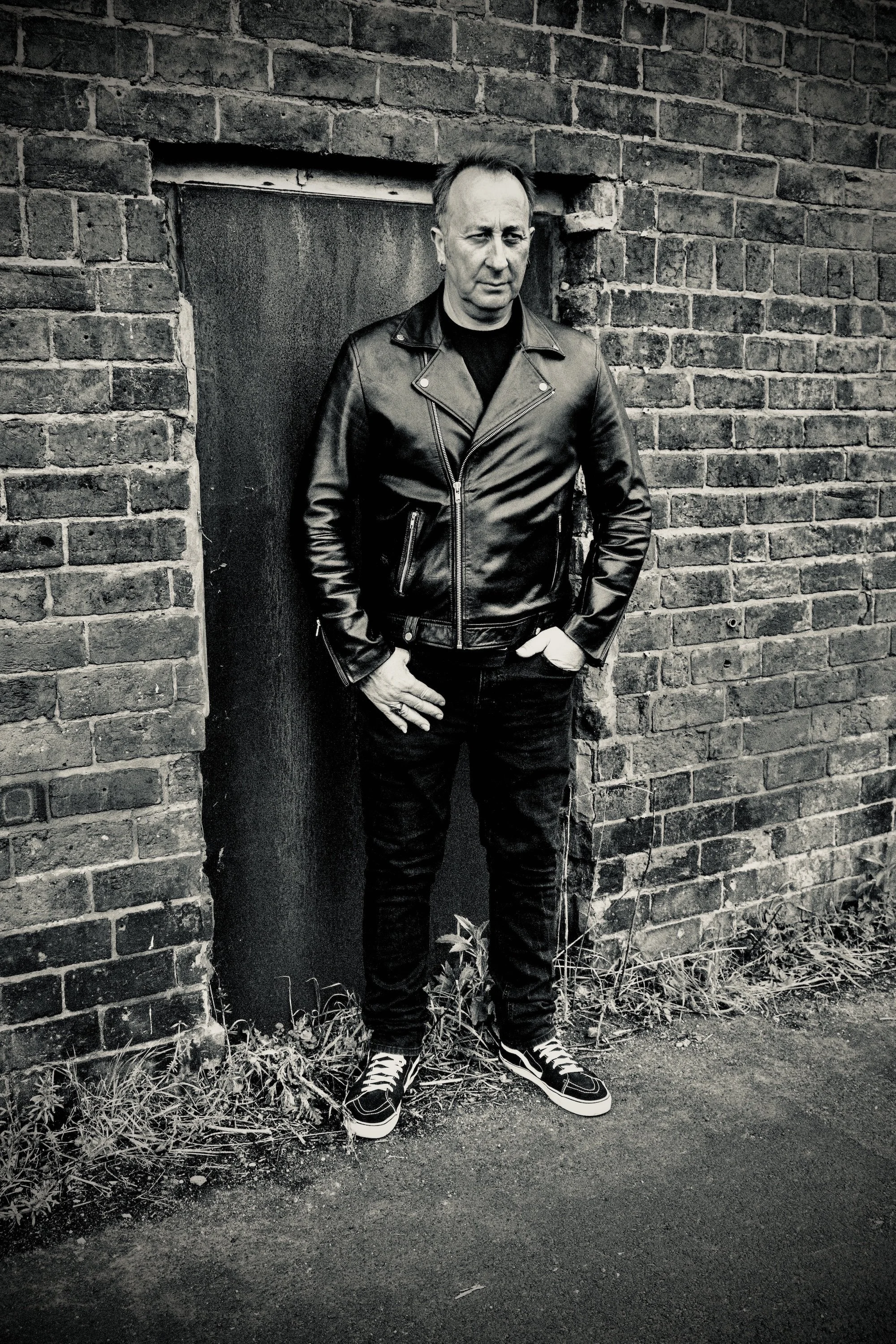 Portrait of a man in a black leather jacket standing before a weathered brick wall — a stark, introspective composition framed by overgrown grass and muted tones.