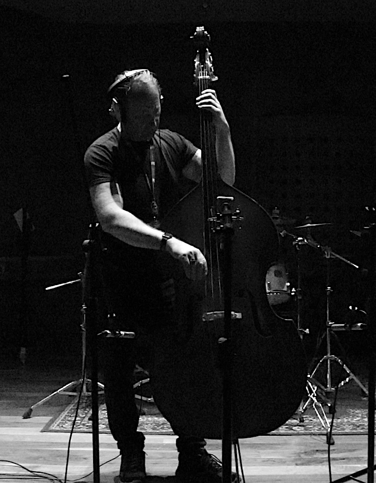 Musician performing on upright bass in a dimly lit studio, headphones on, surrounded by cables and recording equipment — a moment of pure focus.